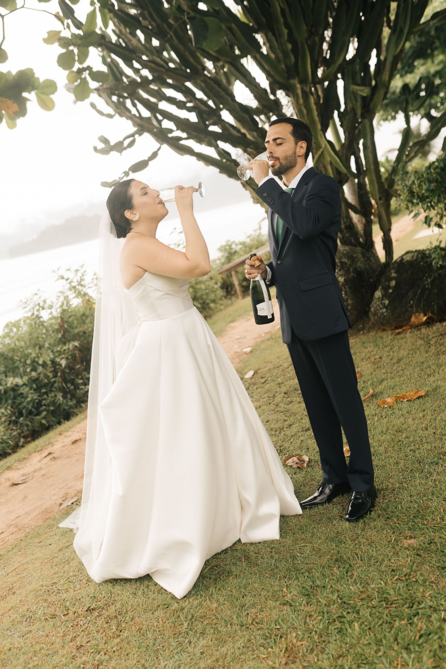 Wedding couple drinking champagne together on a scenic clifftop overlooking the ocean.