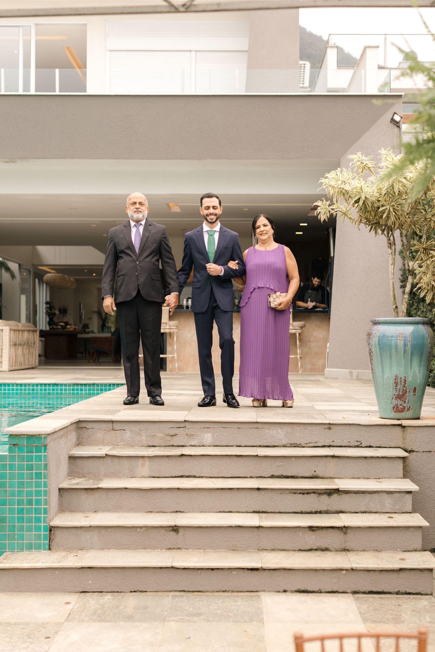 Three guests in formal wear pose on concrete steps by pool.