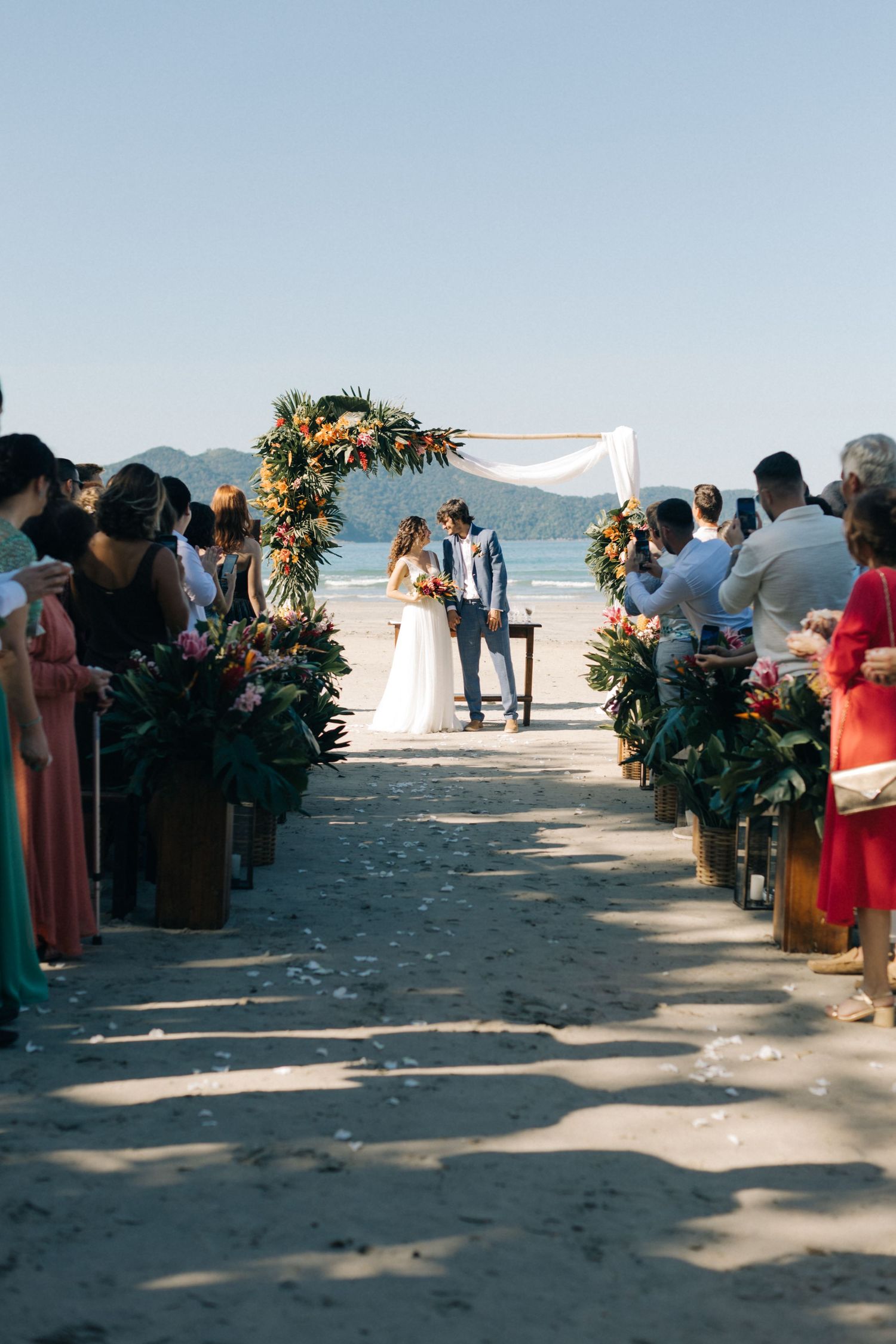 Beach wedding ceremony with floral arch and guests lining sandy aisle.