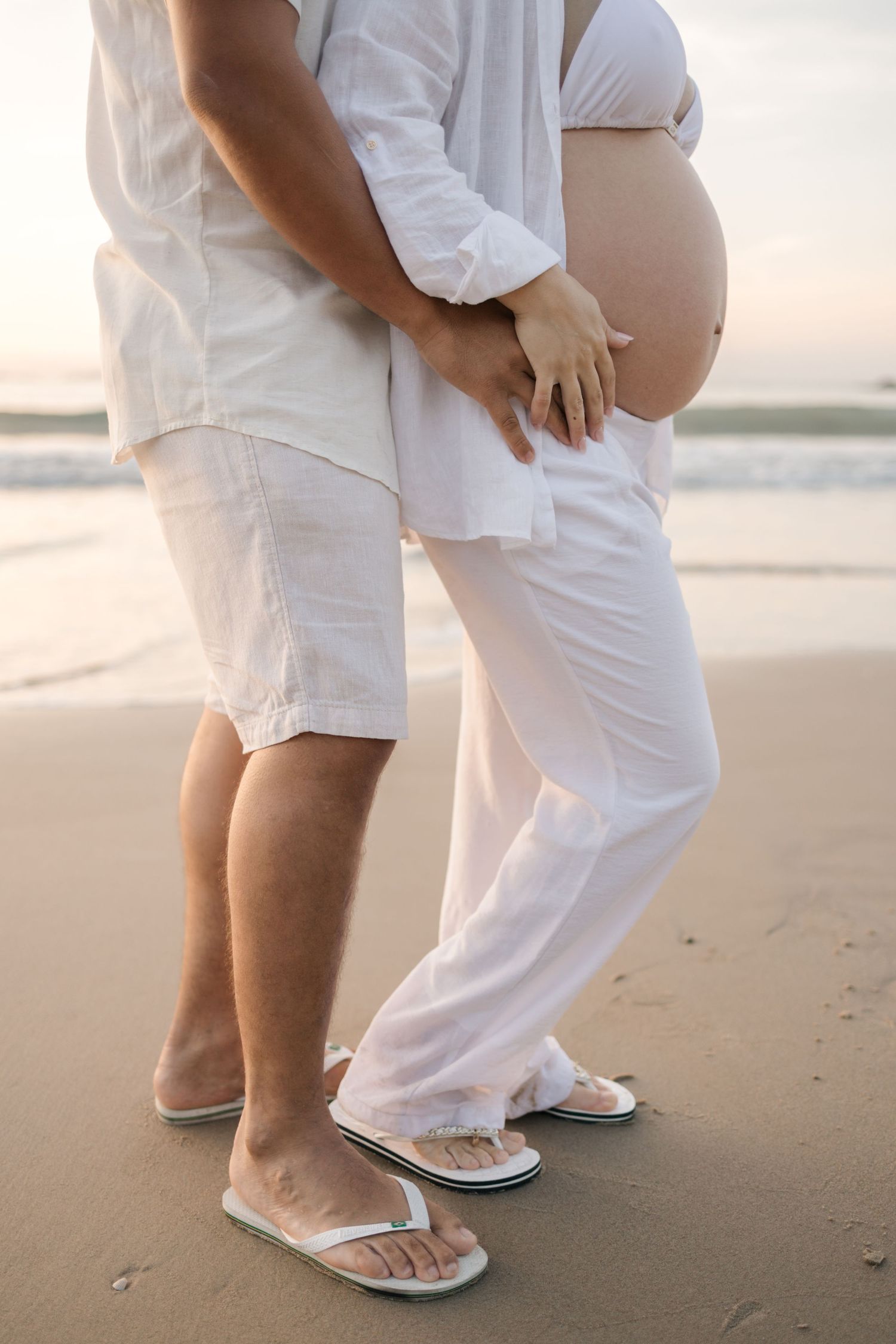 Couple in white beachwear standing close together on sandy shore wearing flip flops.
