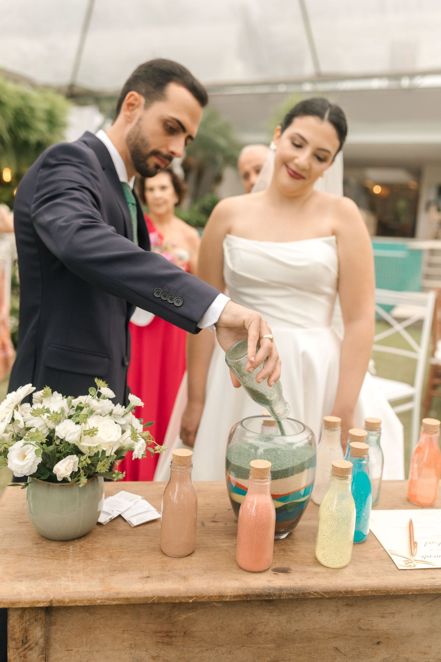 Couple participating in sand pouring ceremony with colorful glass containers on rustic wooden table.