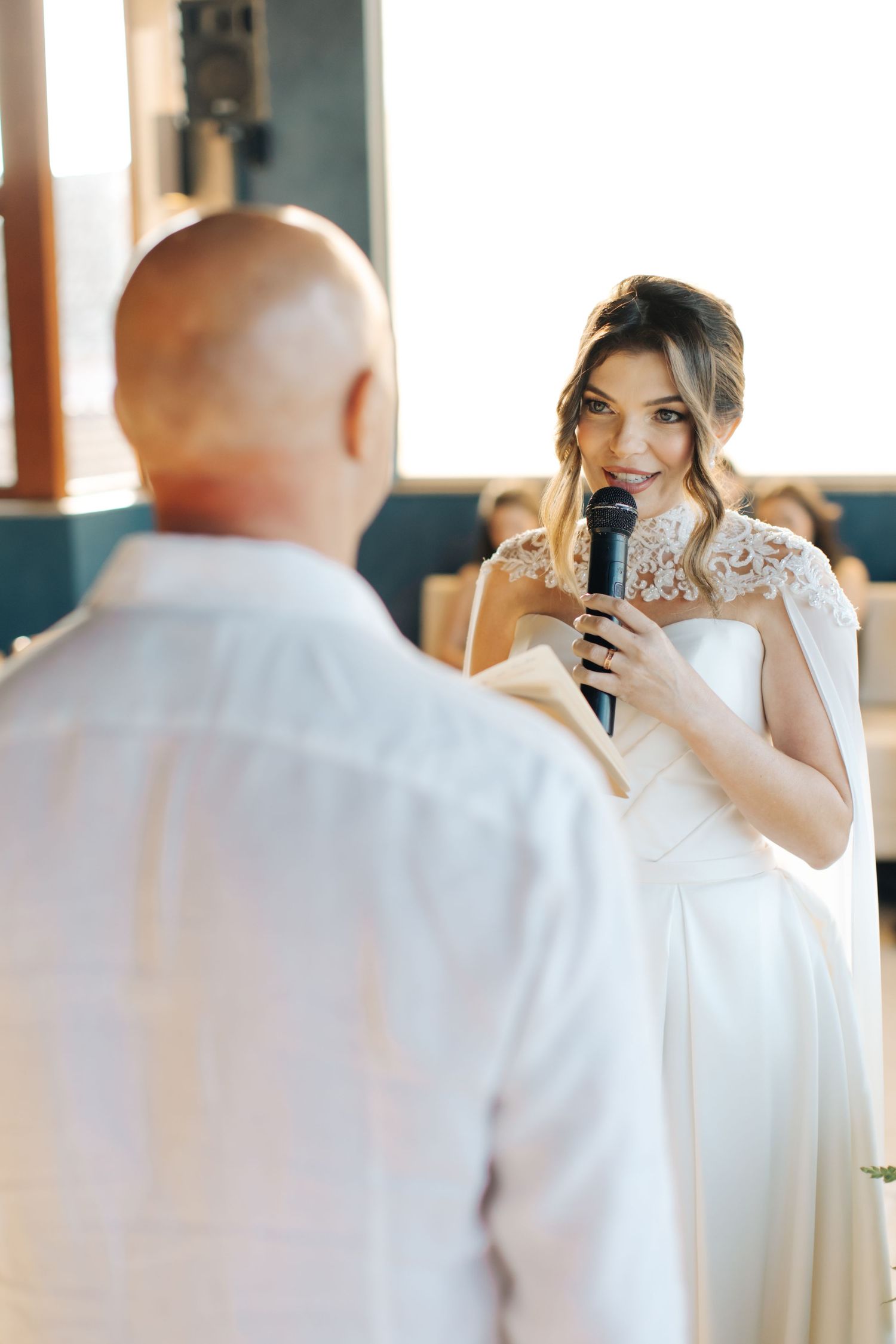 A bride in a white lace dress speaks into a microphone during her wedding ceremony.
