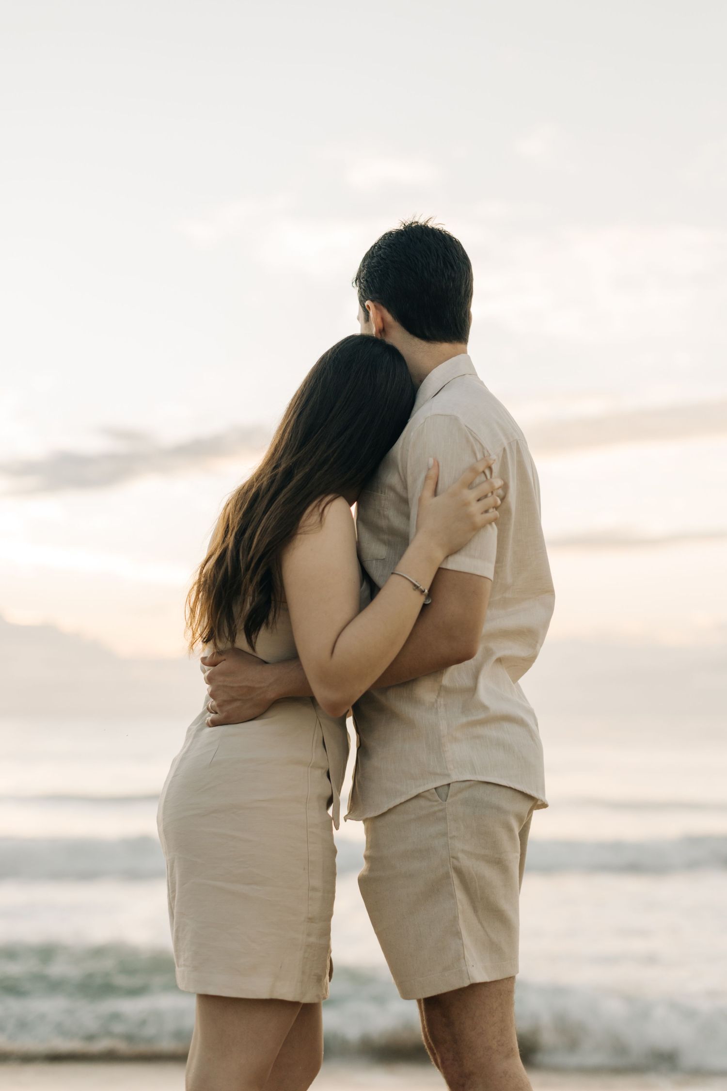 Couple in neutral clothing holding each other close while watching the ocean waves.