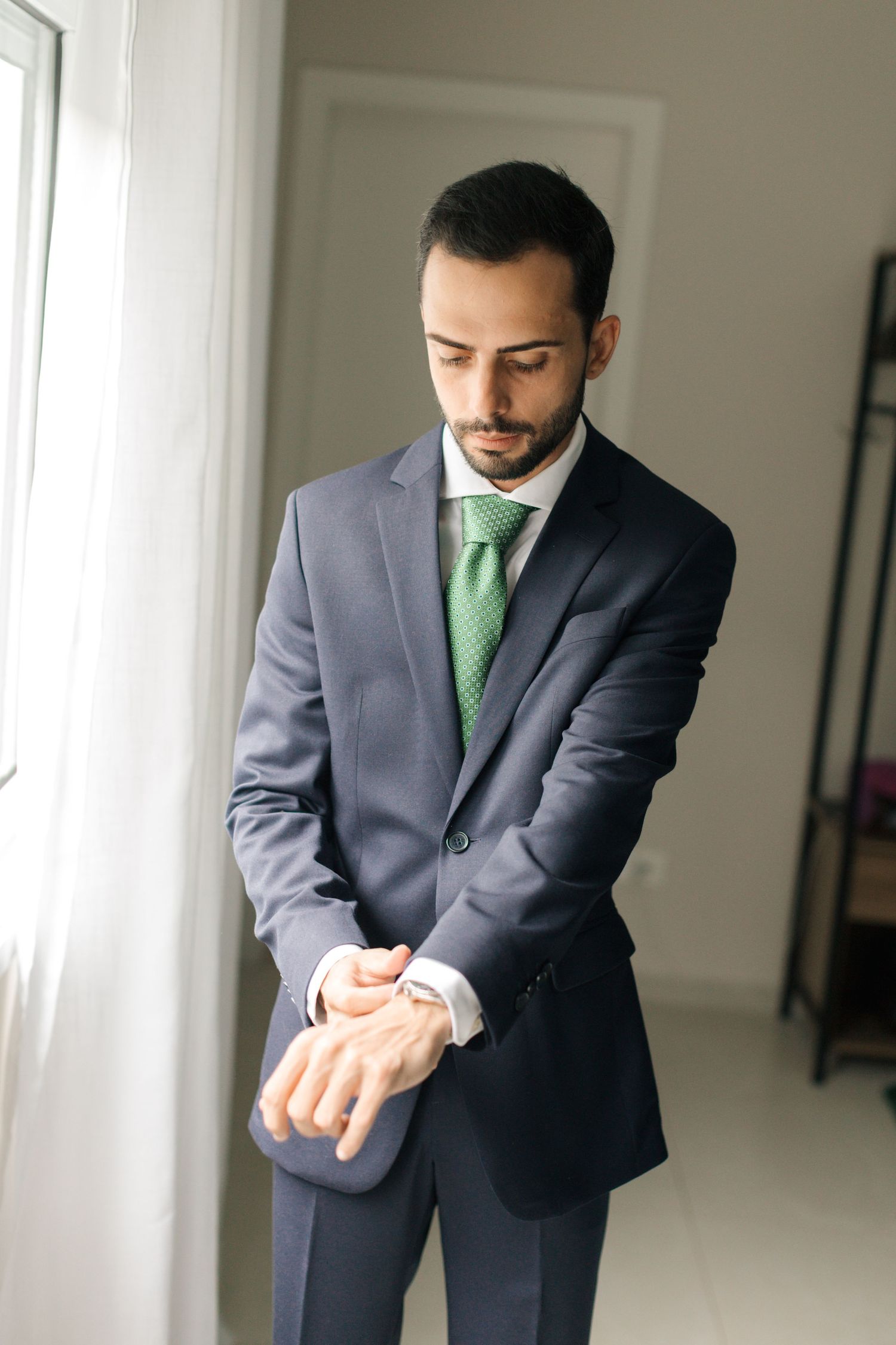 Professional wearing navy suit and green tie checks time on wristwatch near window.