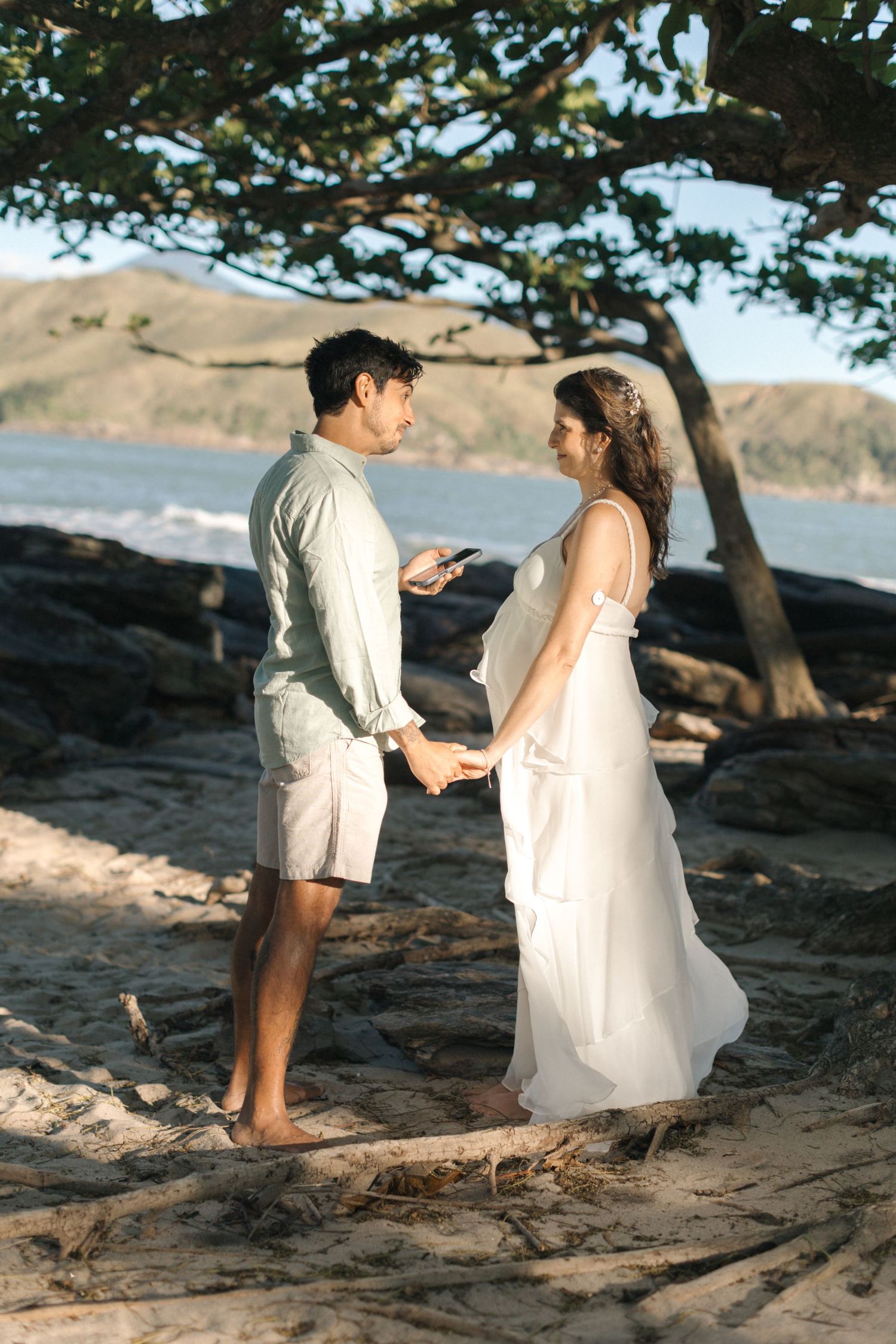 A romantic beach photoshoot at sunset with a couple standing among rocks and trees by the shore.