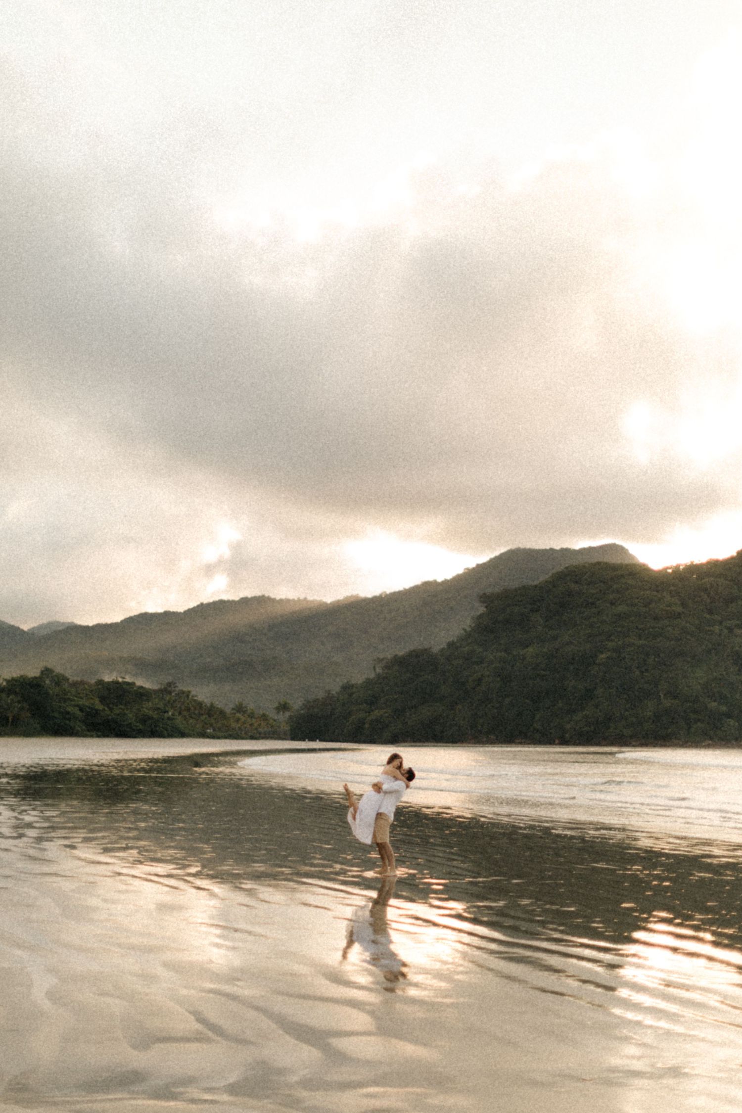 Silhouettes walking on a misty beach with dramatic mountain backdrop at sunset.