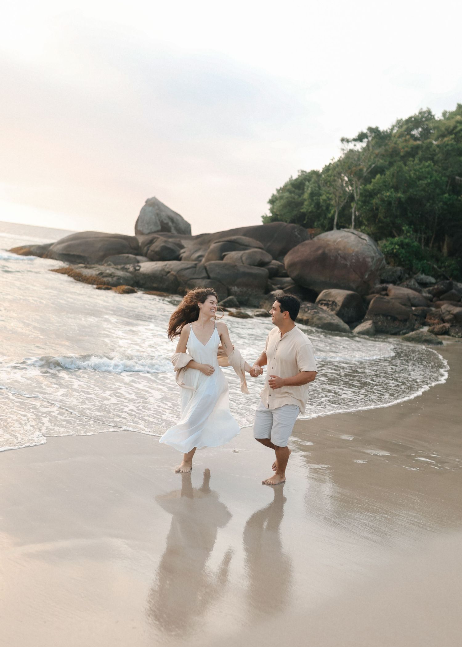 A couple dances joyfully together on a rocky beach at sunset wearing coordinated white outfits.