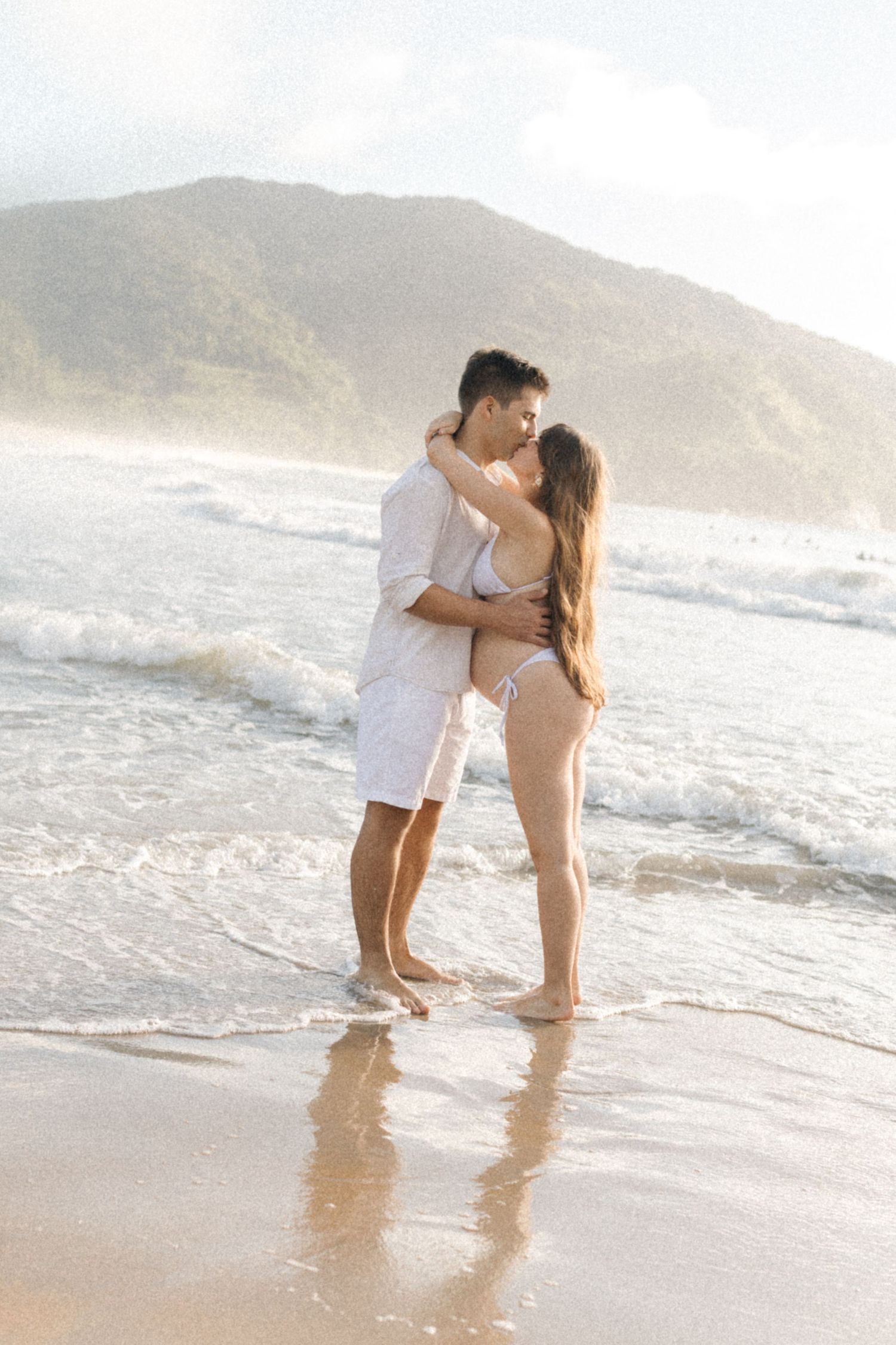 A romantic couple embraces while standing in the ocean waves at sunset on a misty beach.