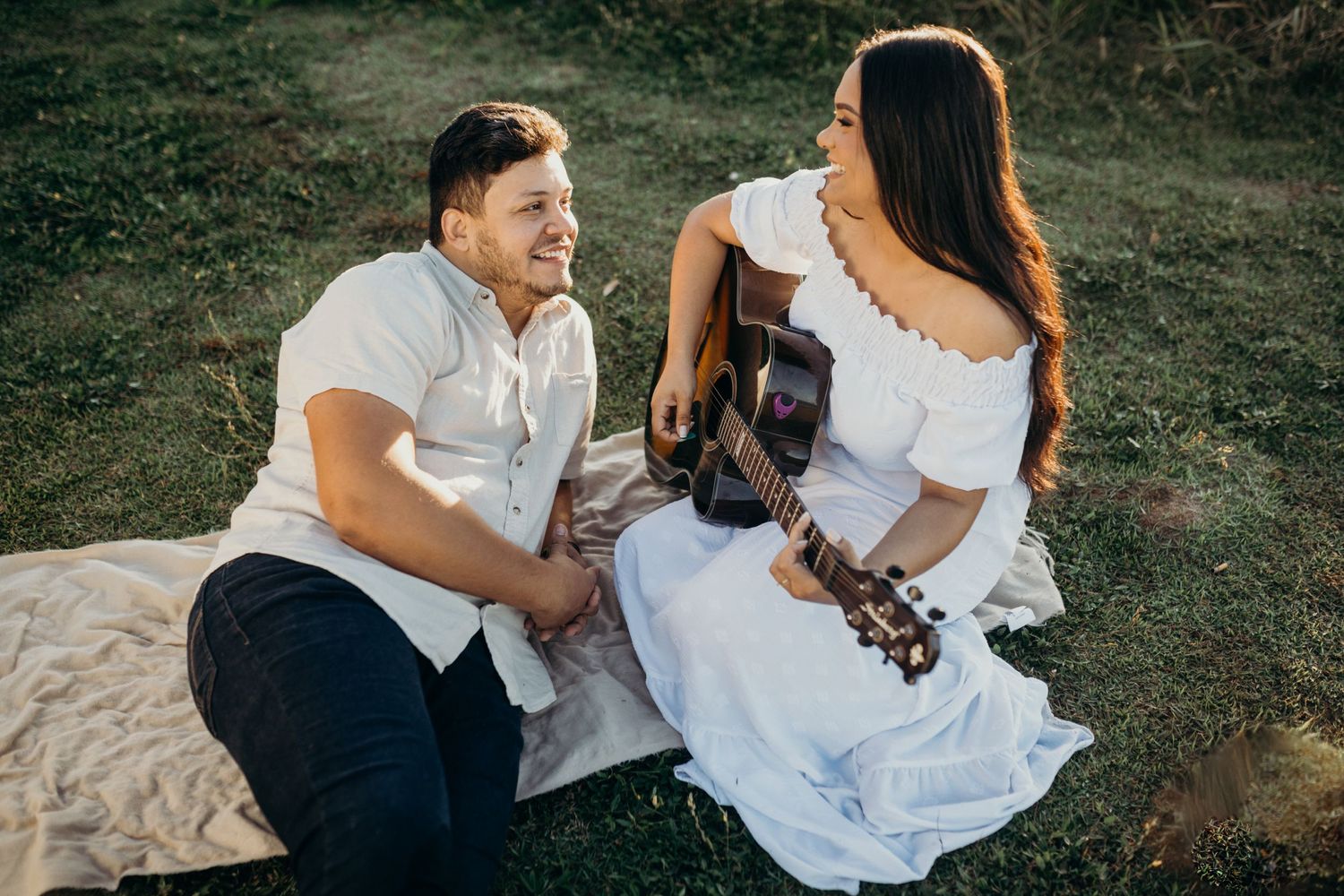 Couple shares joyful moment with guitar while sitting on picnic blanket in nature.