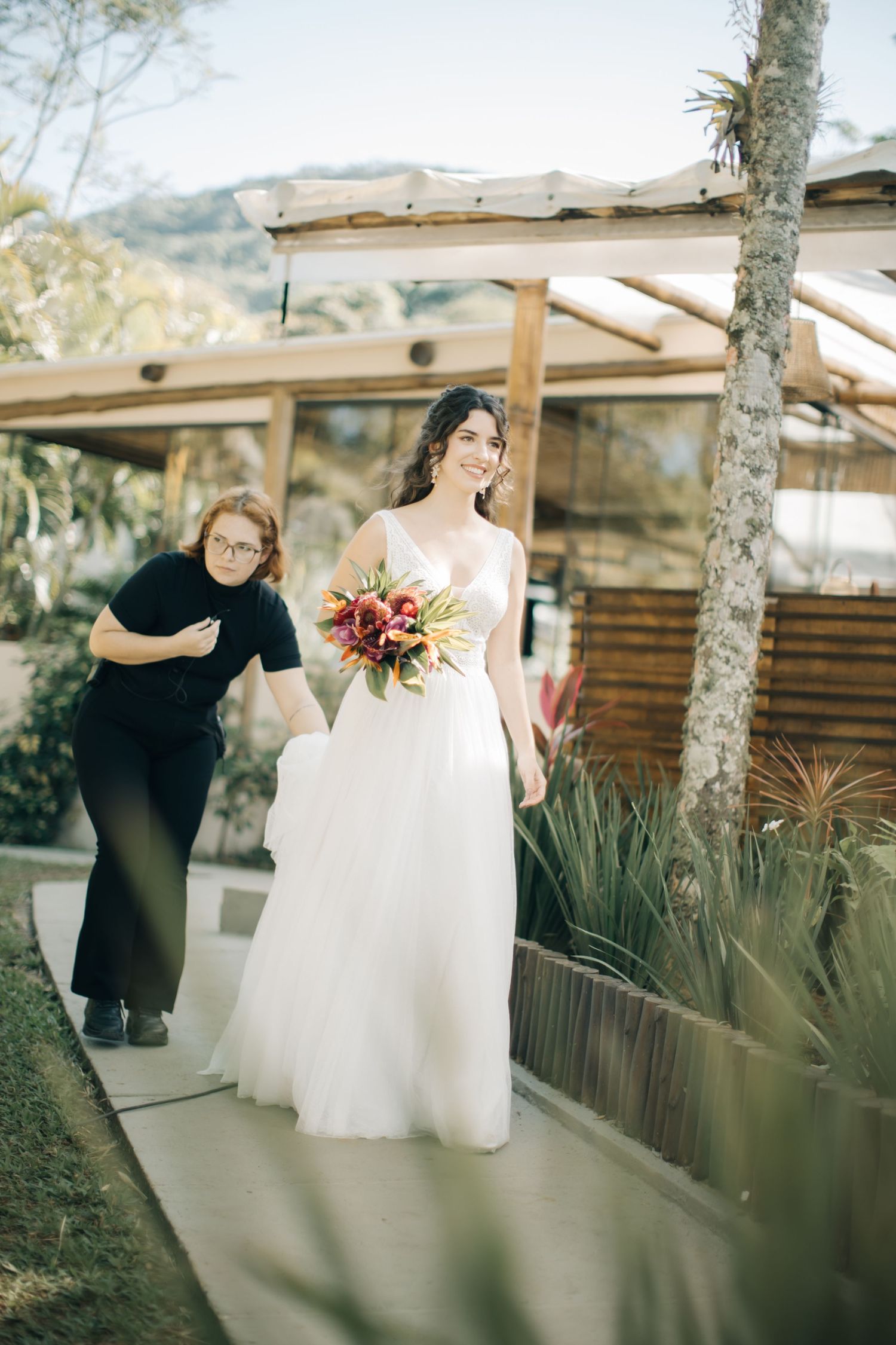 A bride in a flowing white gown carries a tropical bouquet along an outdoor pathway.