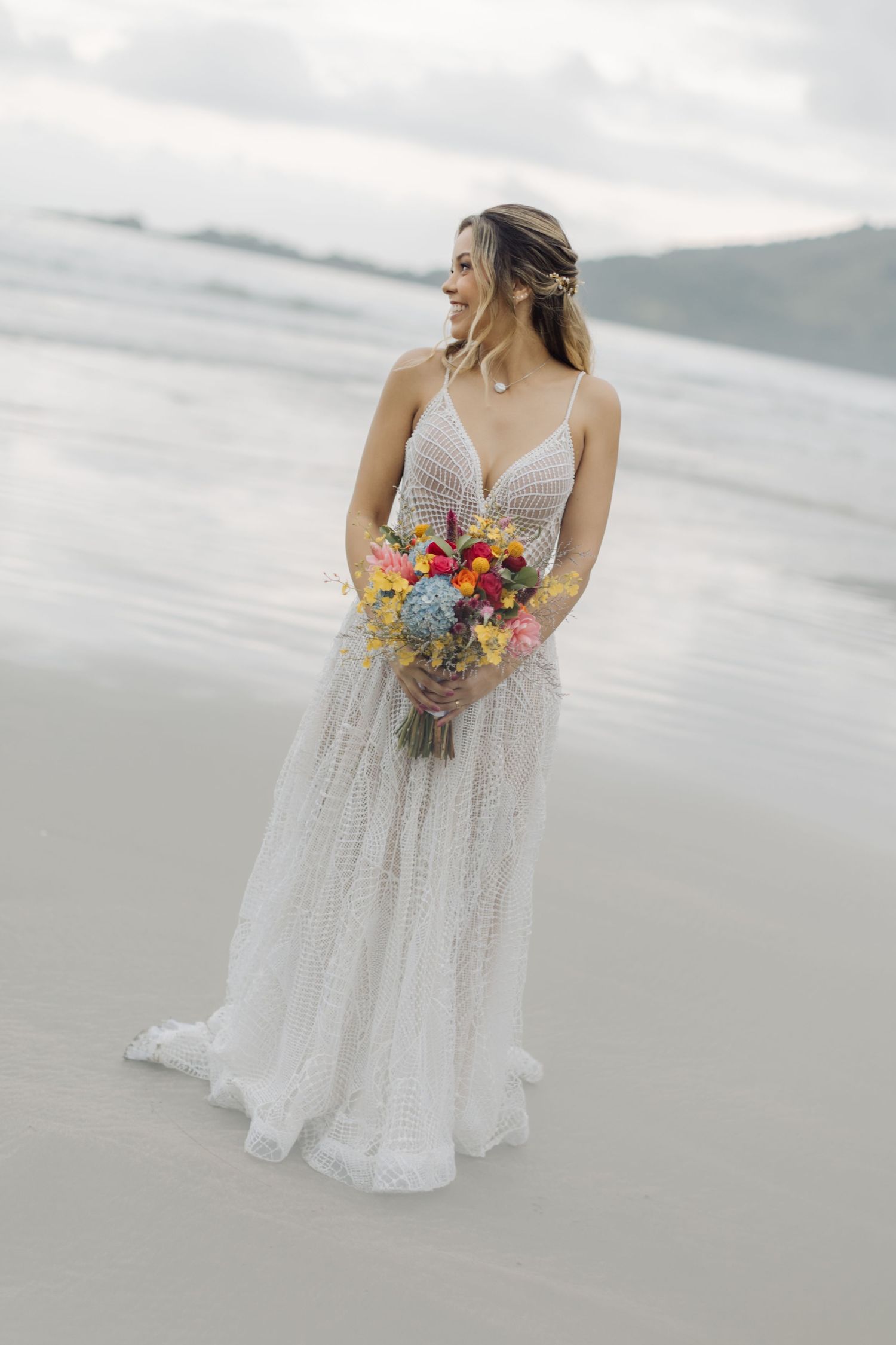 A bride poses with her vibrant floral bouquet against a cloudy oceanfront backdrop.