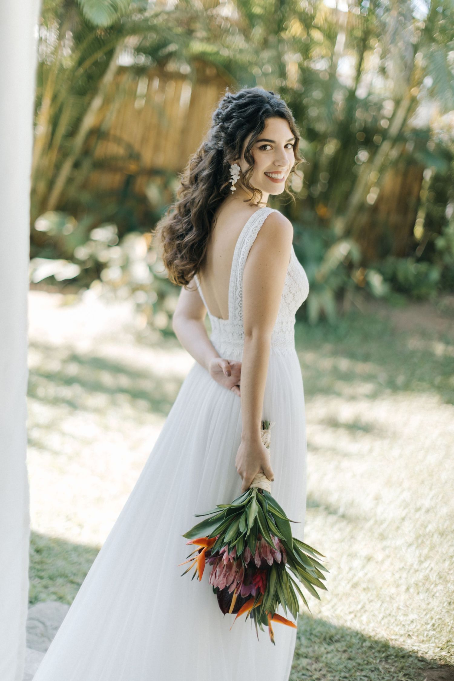 Bride in empire waist dress holds colorful tropical bouquet on sunny porch.