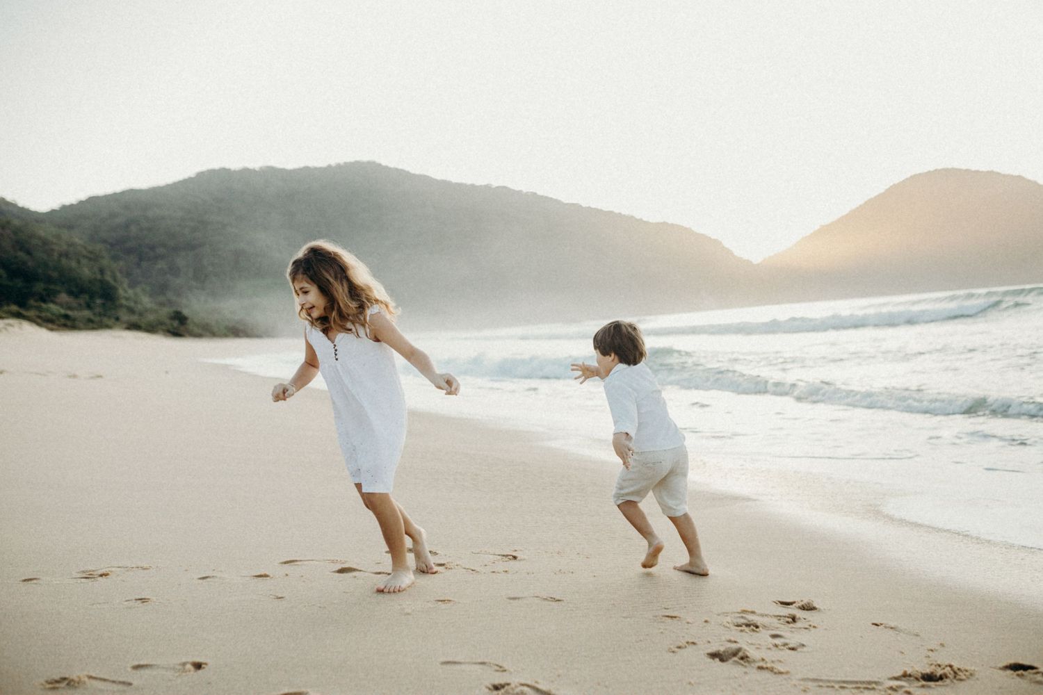 Two silhouettes playing and running on a beach at sunset with mountains in background.