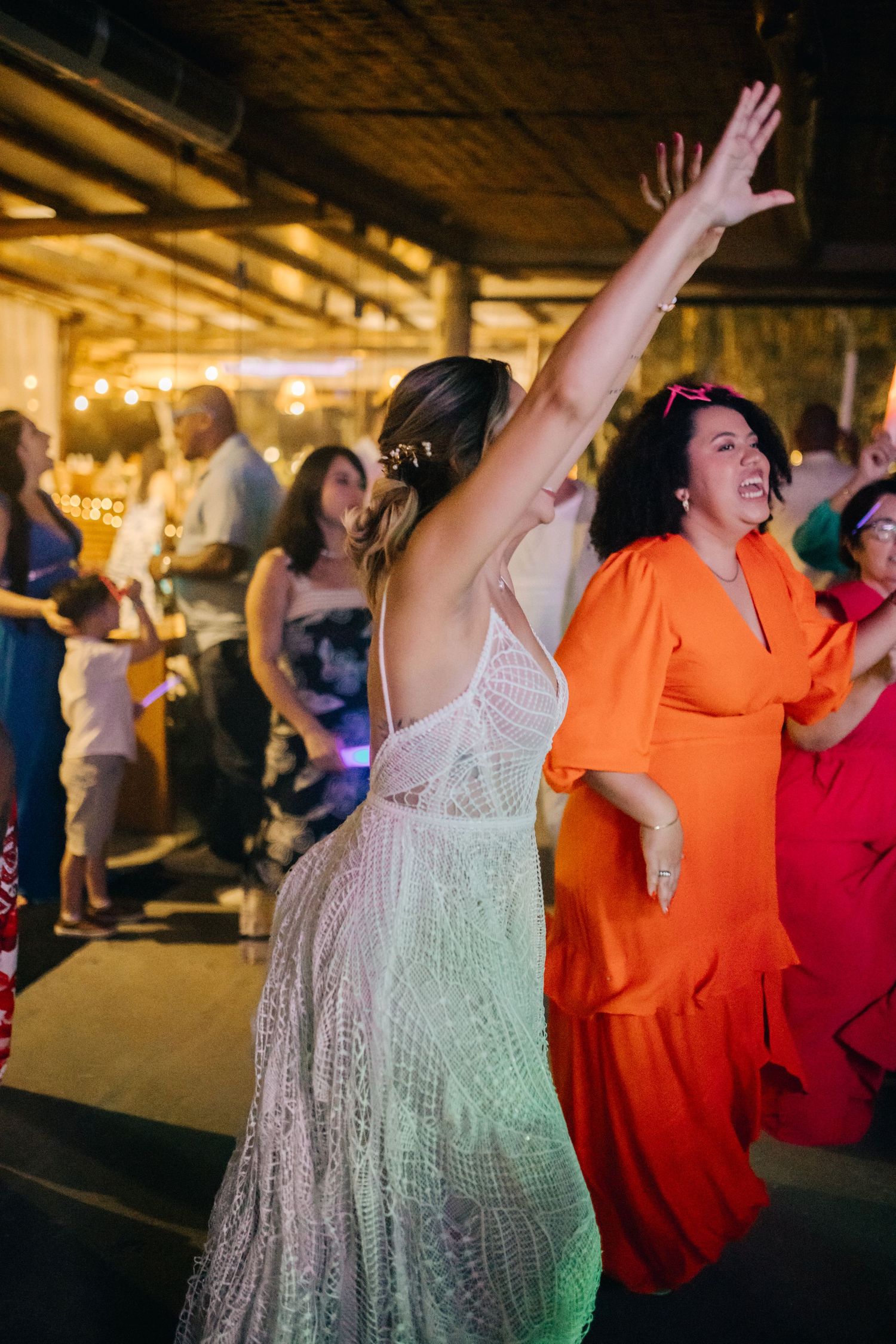 Wedding guests in colorful dresses dancing with raised arms at a reception celebration.