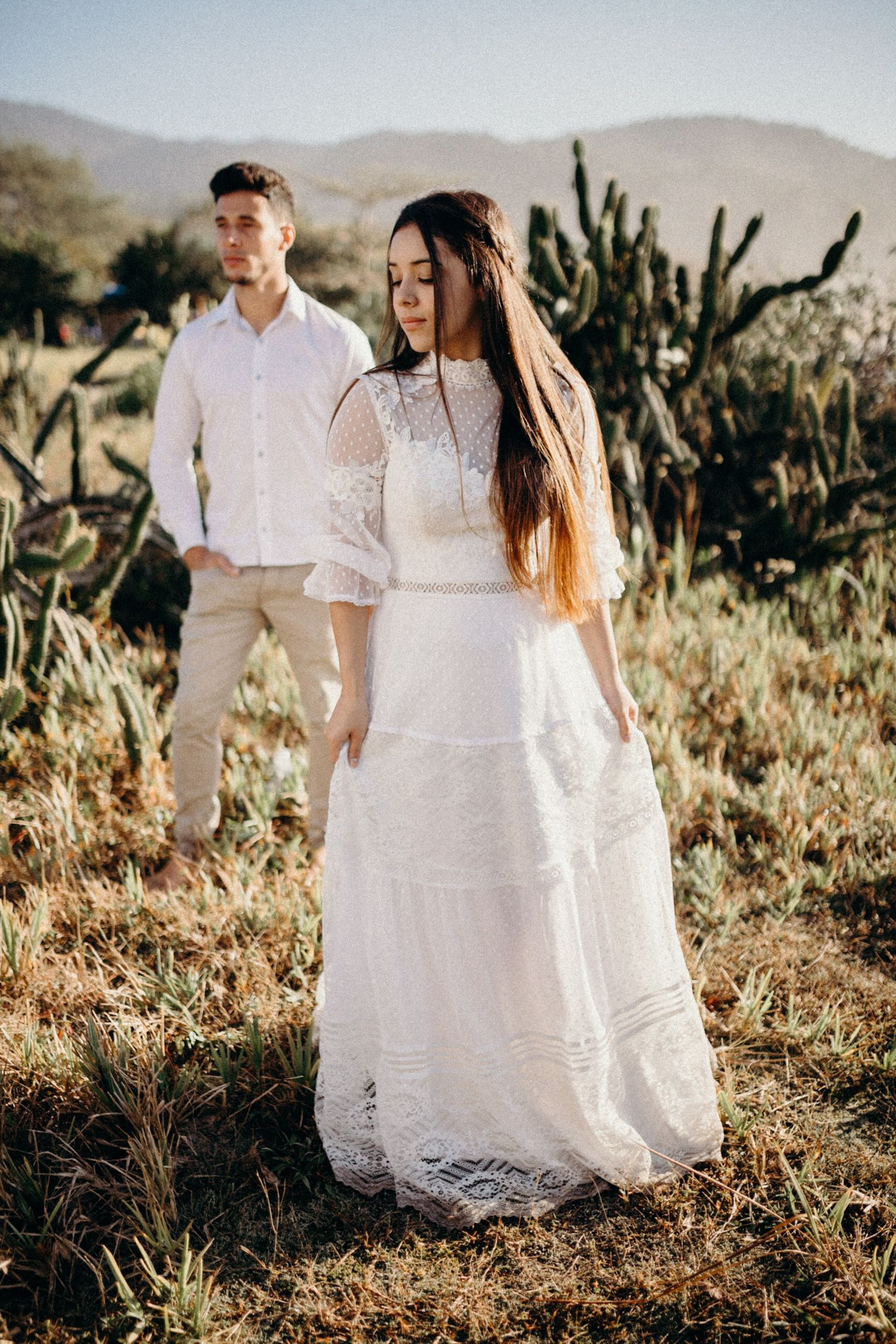 Couple in white attire pose against desert mountain backdrop with saguaro cacti.