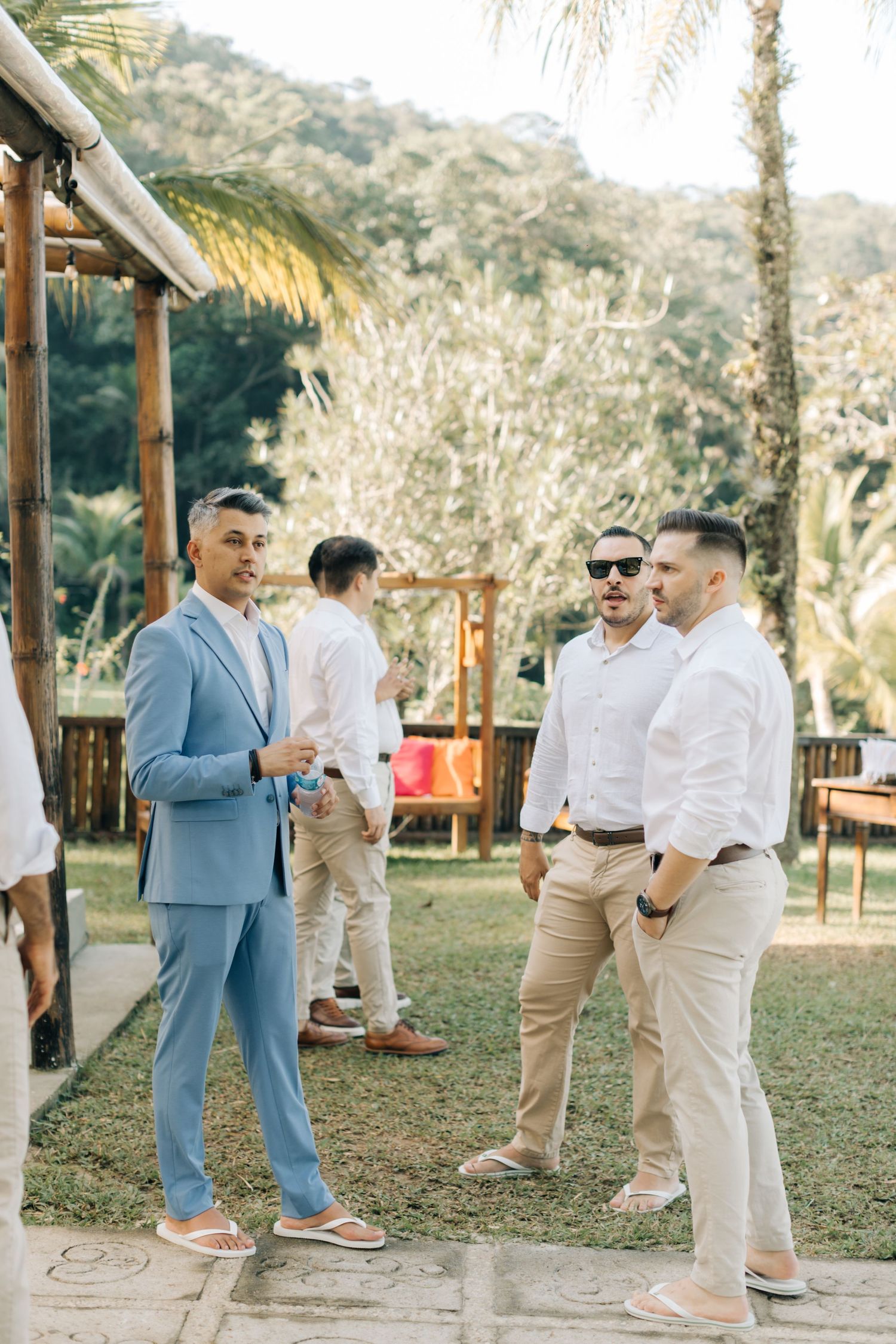 Wedding guests in light colored suits mingle on a palm-tree lined outdoor patio.