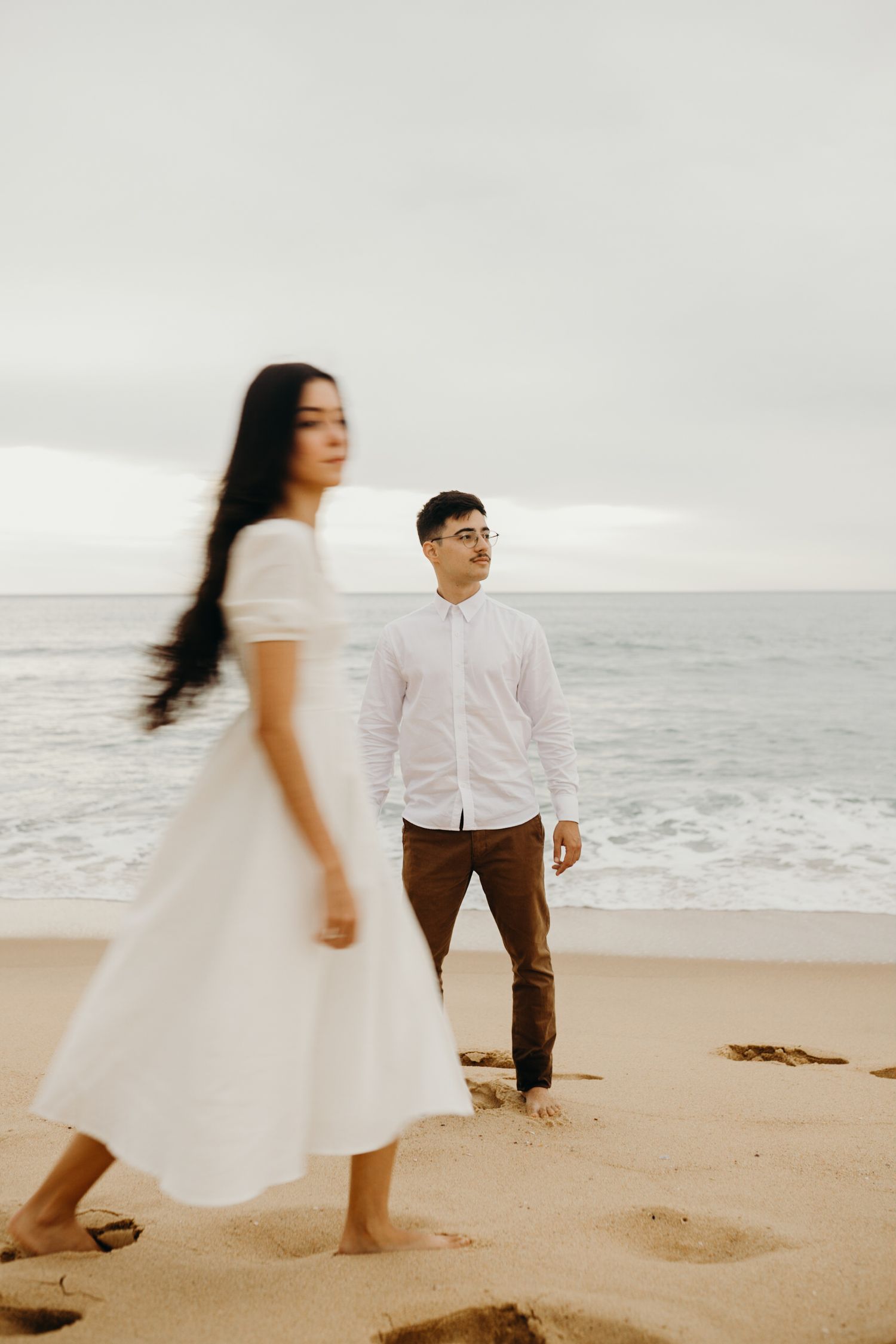 A romantic couple in white outfits standing on a beach at sunset with ocean waves in the background.