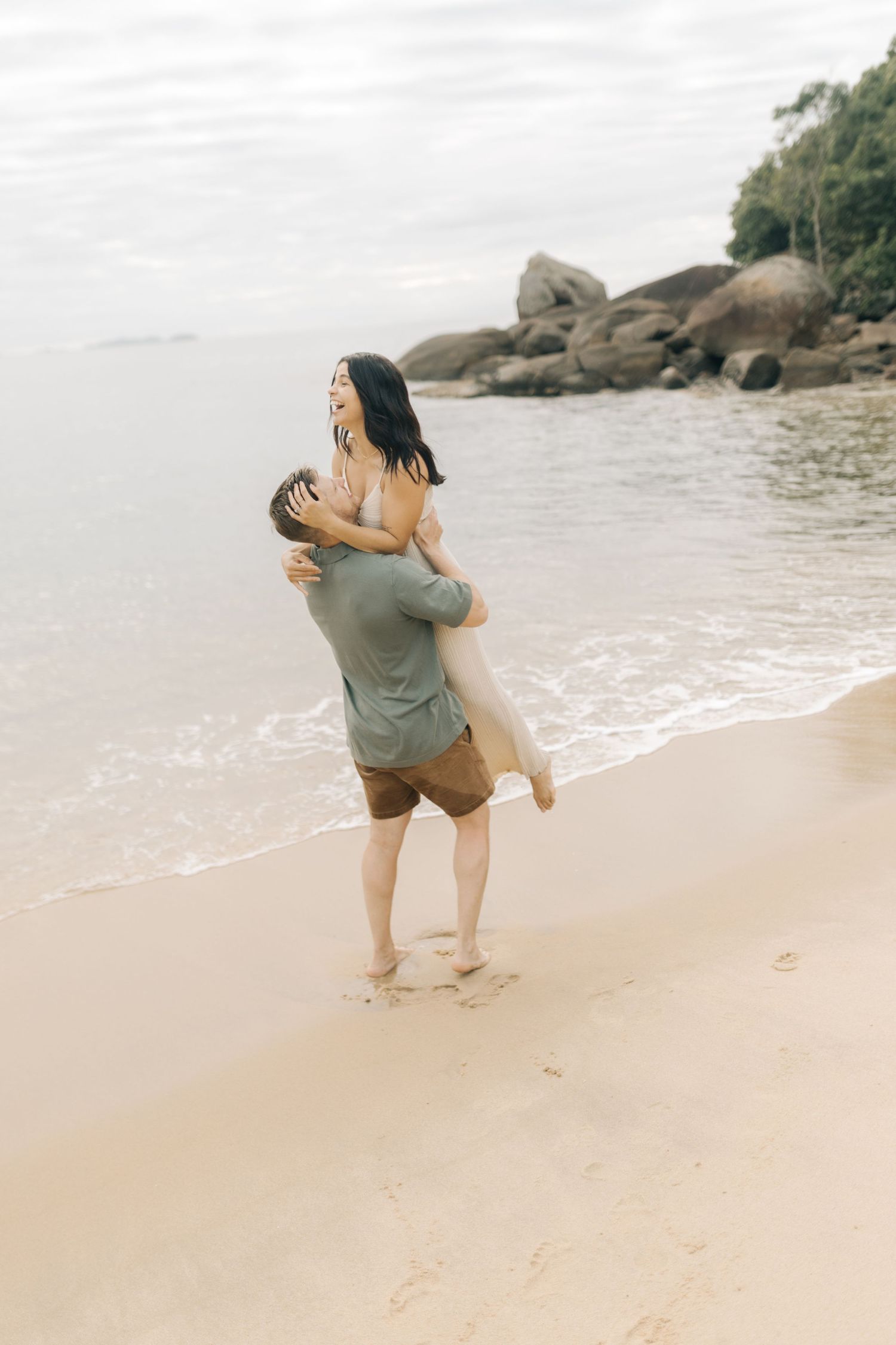 Two people share a joyful embrace on a secluded beach with large boulders in the background.