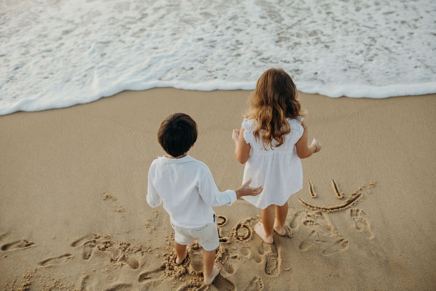 Two children making sand art on the beach as waves roll in during sunset.