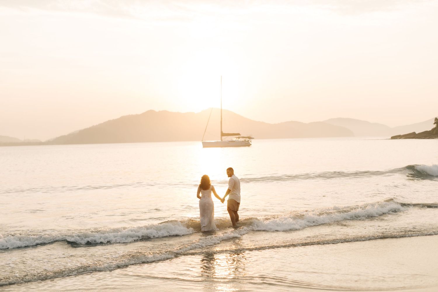 A couple stands in gentle waves at sunset with a sailboat and mountains in the background.