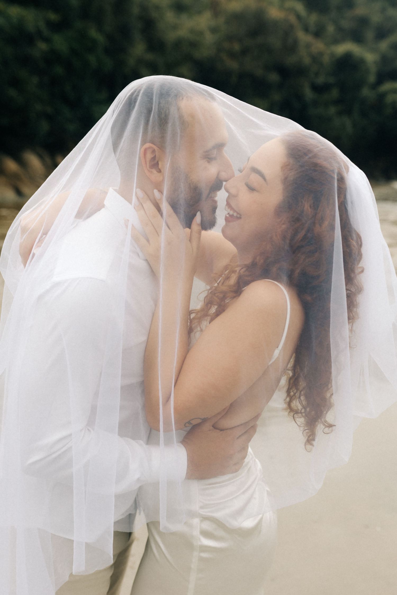 Romantic couple sharing tender moments under a flowing white veil on a beach.
