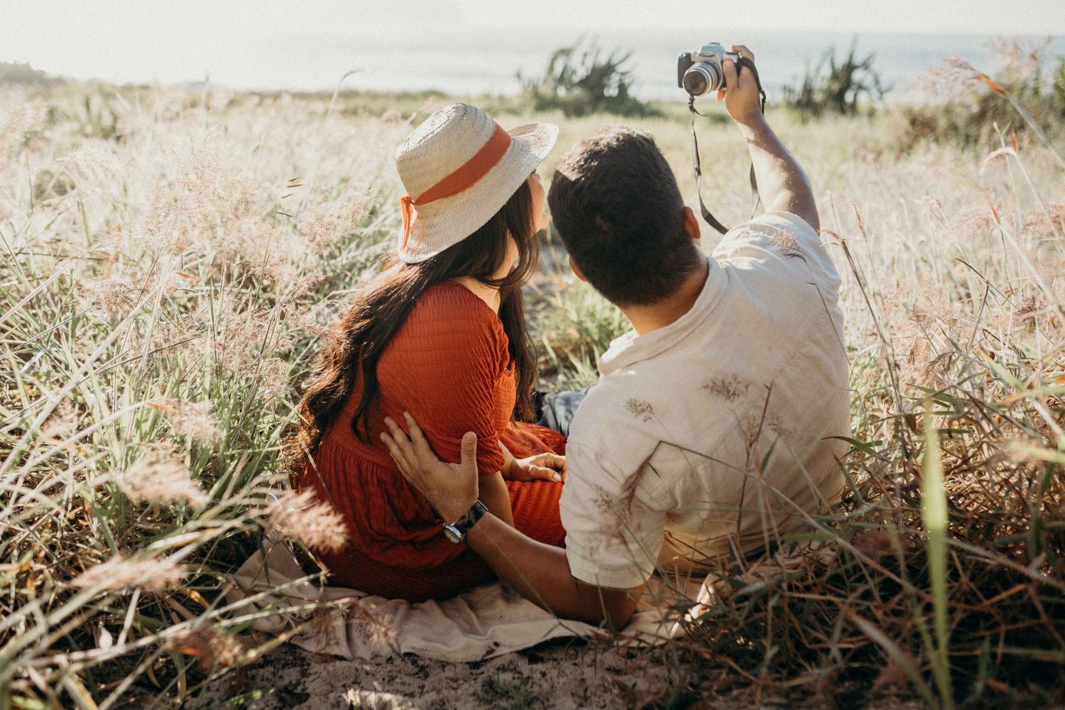 A couple sits together in golden grass overlooking the ocean while taking photographs.
