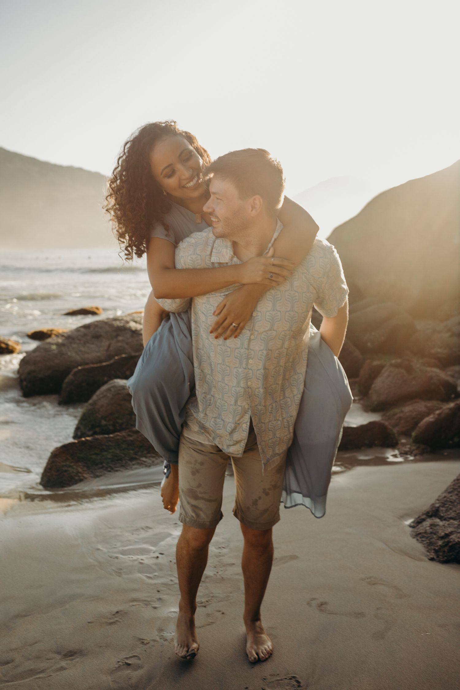 Couple sharing joyful moments on a beach with rocky coastline at golden hour.