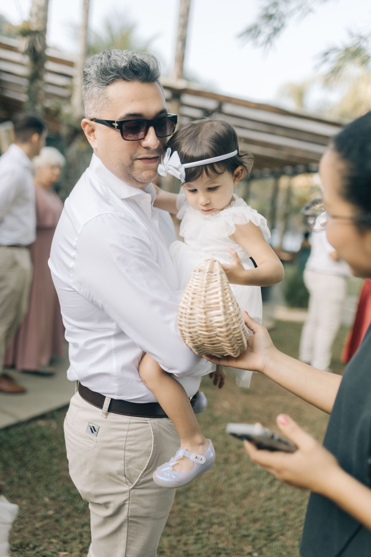 A small child in white dress being held at an outdoor wedding celebration with a wicker basket.