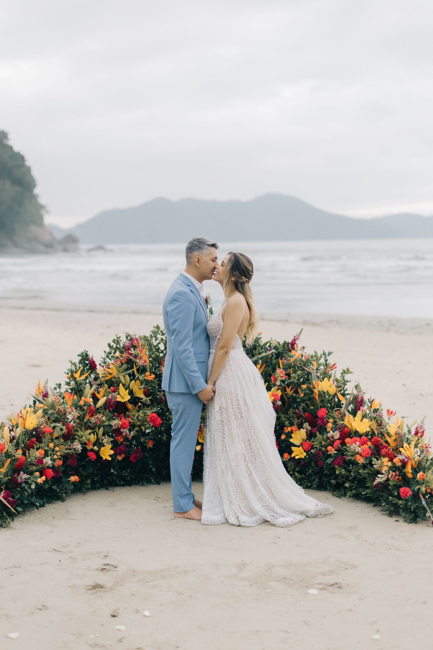 A romantic beach wedding ceremony with a couple embracing in front of a vibrant floral arch by the ocean.
