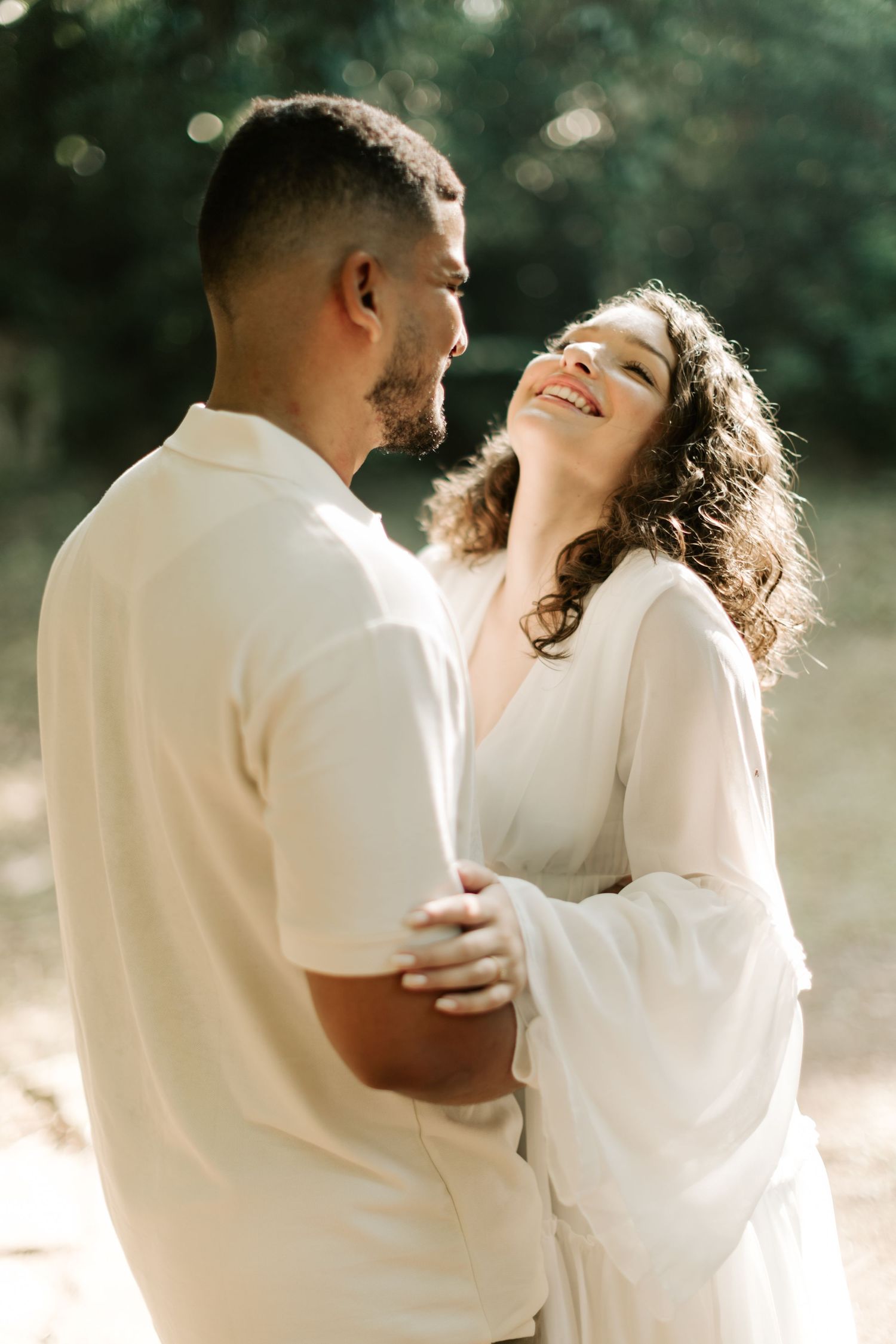 A couple in white outfits shares a tender moment in dappled sunlight outdoors.