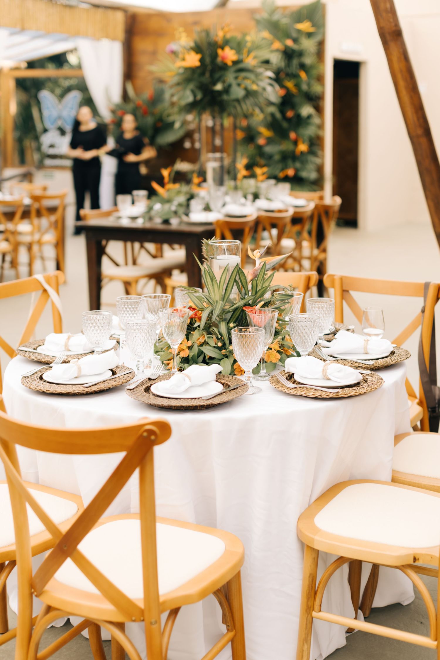 Round dining table with white tablecloth, wooden chairs and tropical centerpiece in natural light setting.
