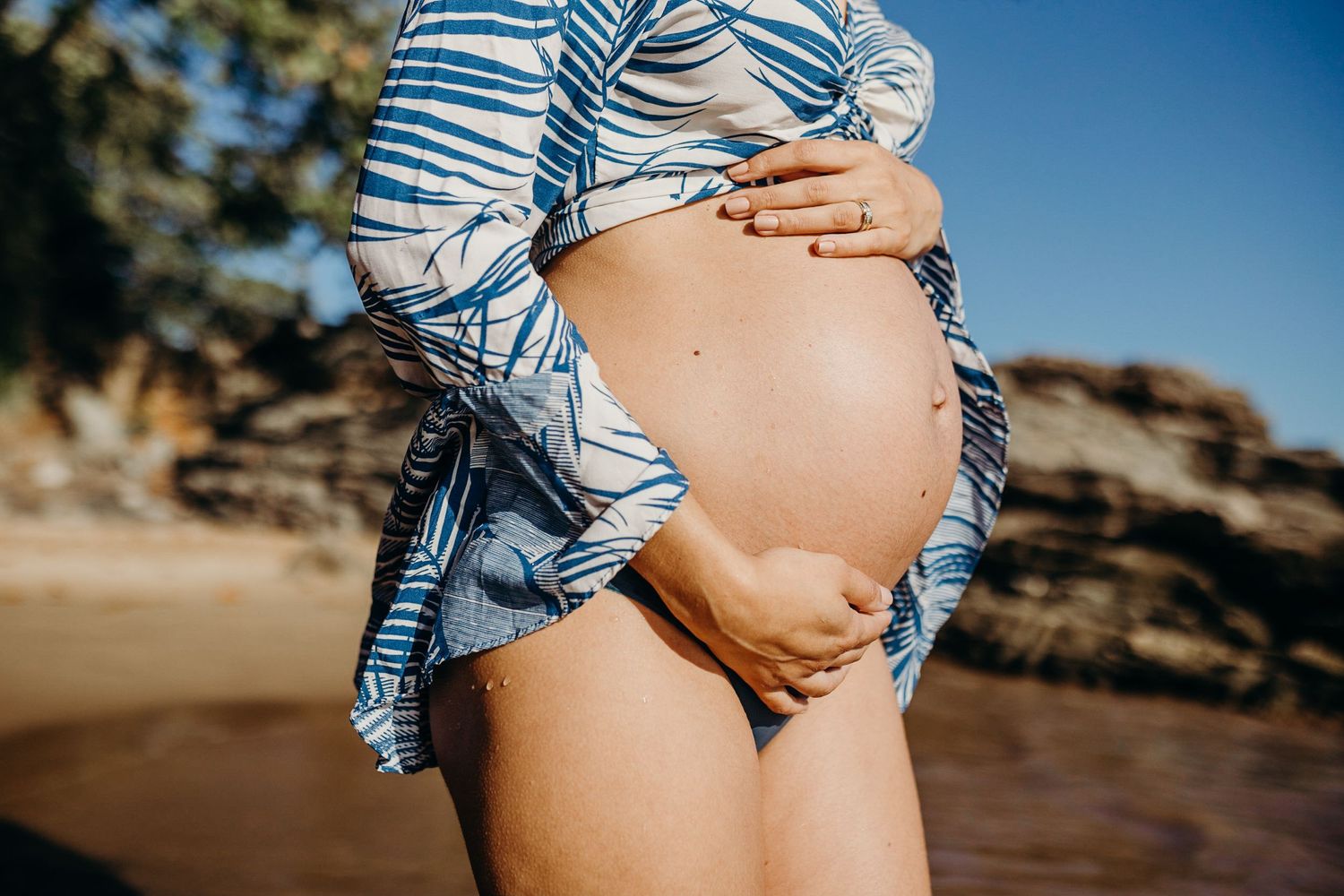 Close-up shot of a pregnant belly with a blue and white patterned cover-up on a beach.