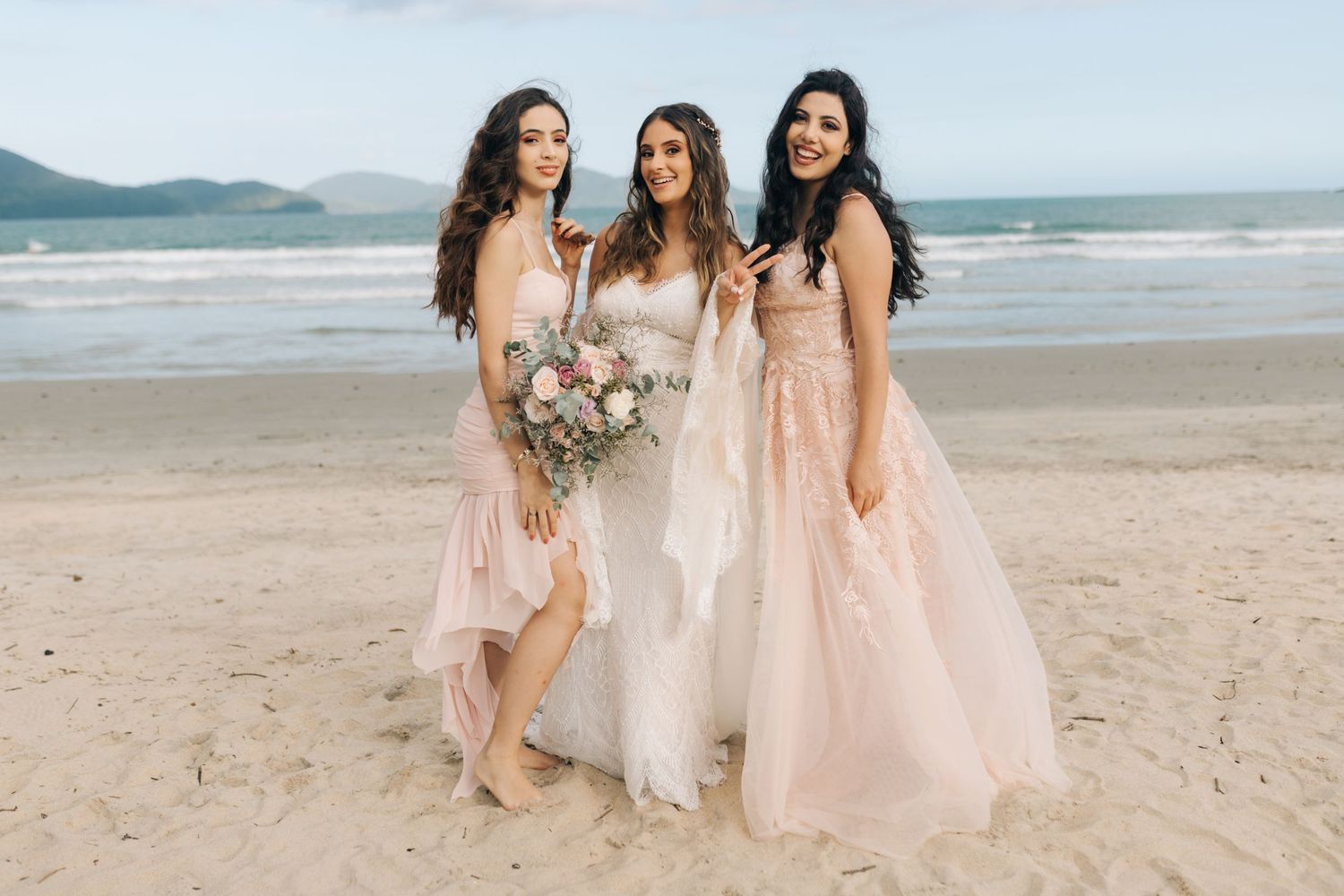 Bridesmaids in flowing pink dresses pose with bride holding elegant bouquet on sandy beach.
