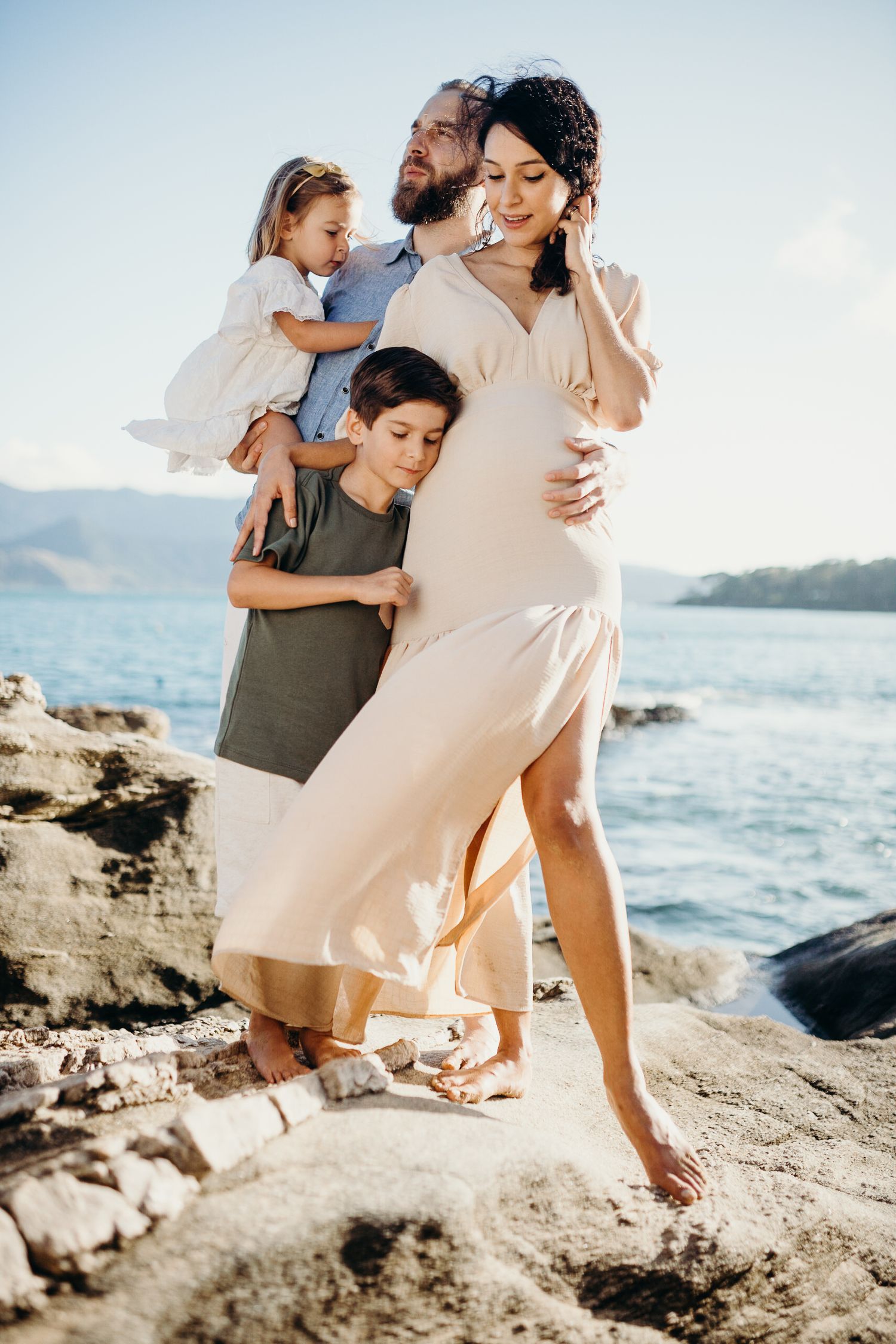 Family embraces expectant mother in flowing dress on rocky beach with ocean and mountains in background.