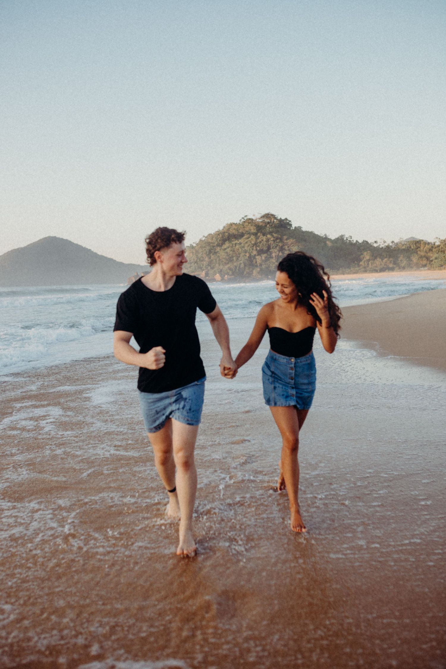 Happy couple runs together along beach shoreline at sunset with mountains in background.