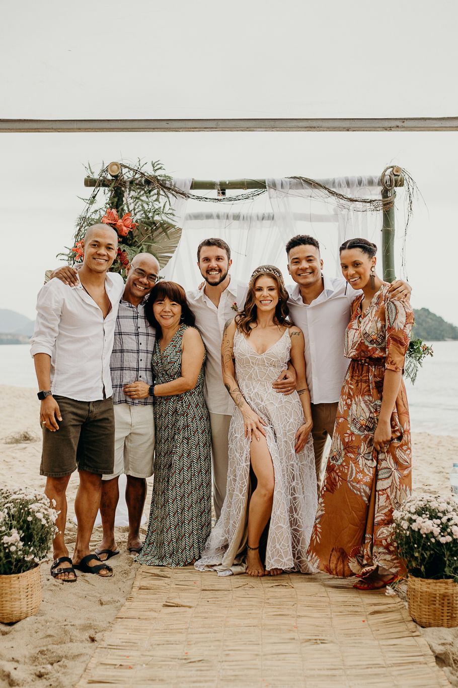 A group photo of wedding guests under a decorated arch at a scenic beach location.