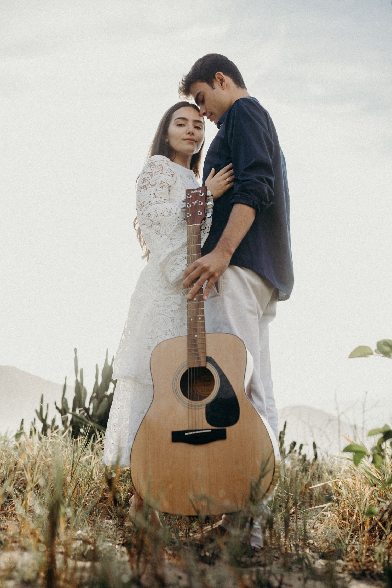 A romantic outdoor portrait featuring an acoustic guitar and flowing white lace dress against tall grass.