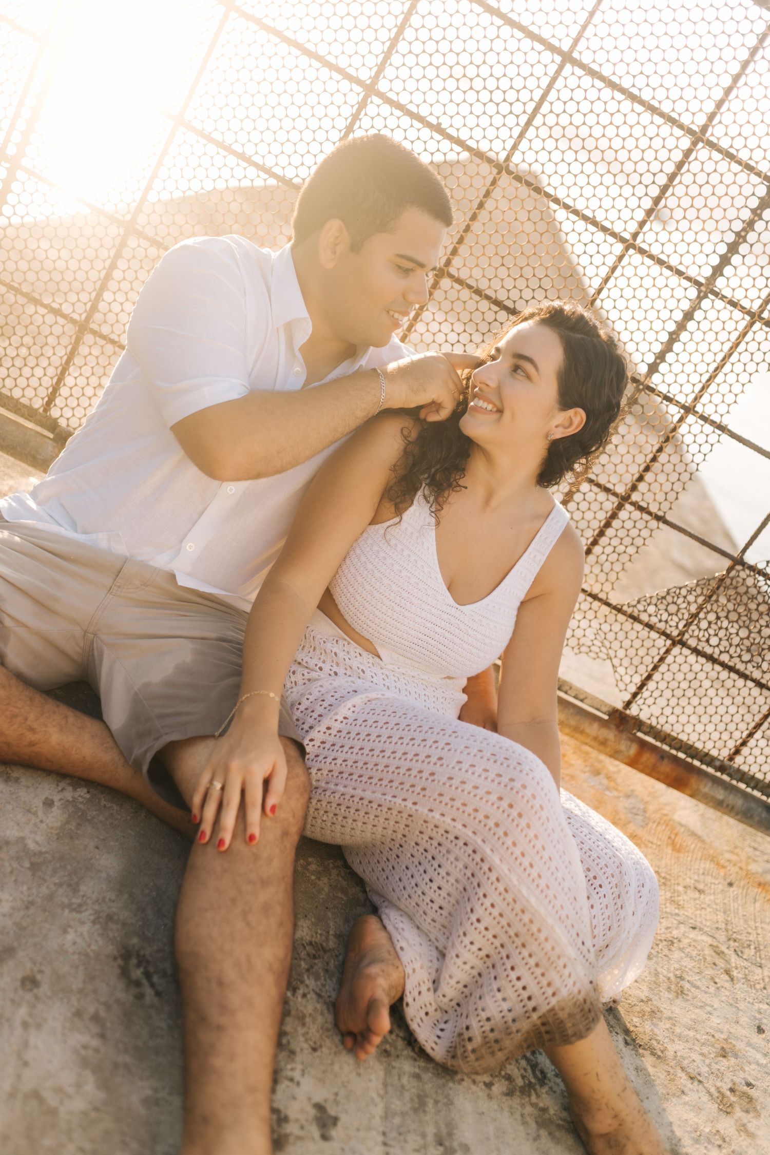 A couple sharing a playful moment while sitting on concrete with chain link fence background.
