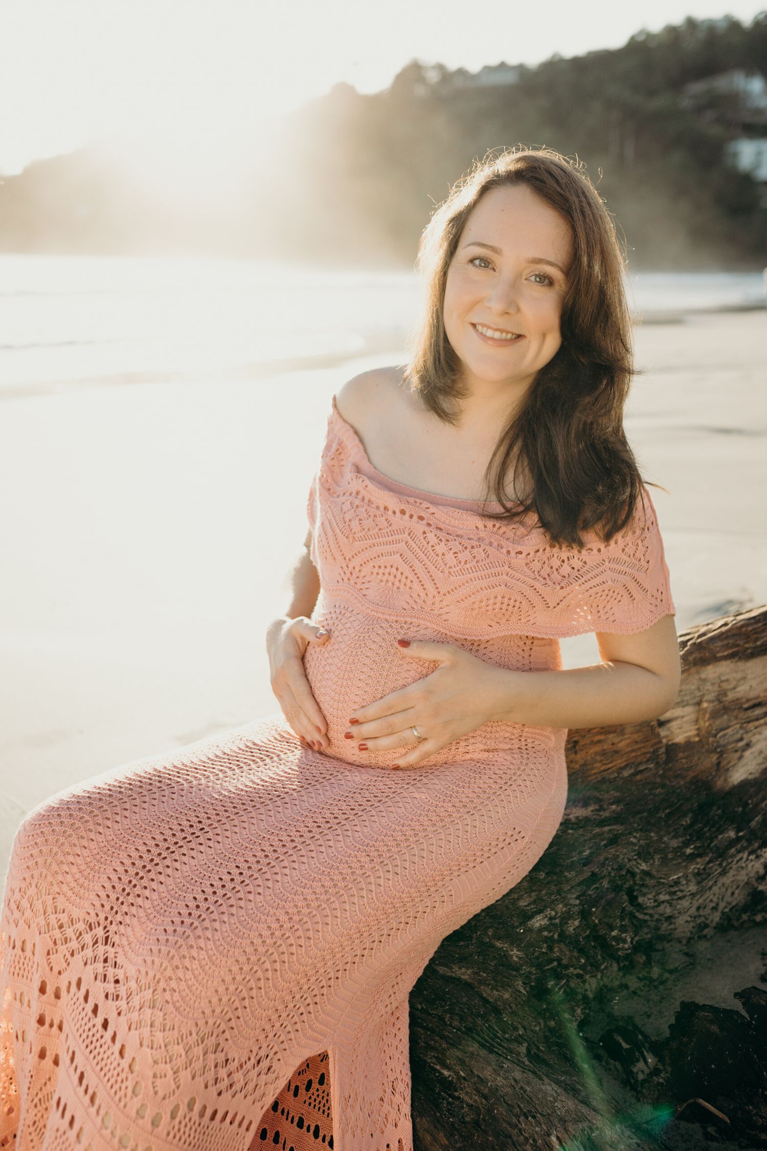 Maternity photos of woman in peach dress sitting on beach rocks at golden hour.