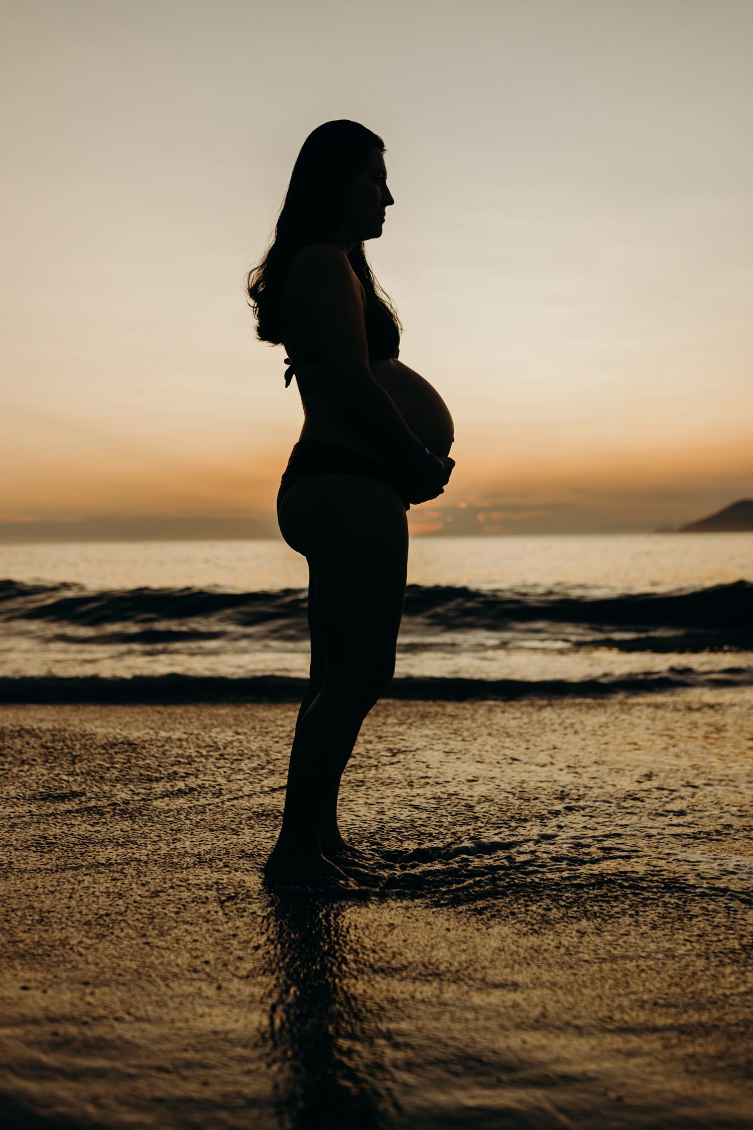 Silhouette of an expectant mother standing on the beach at sunset highlighting her baby bump.