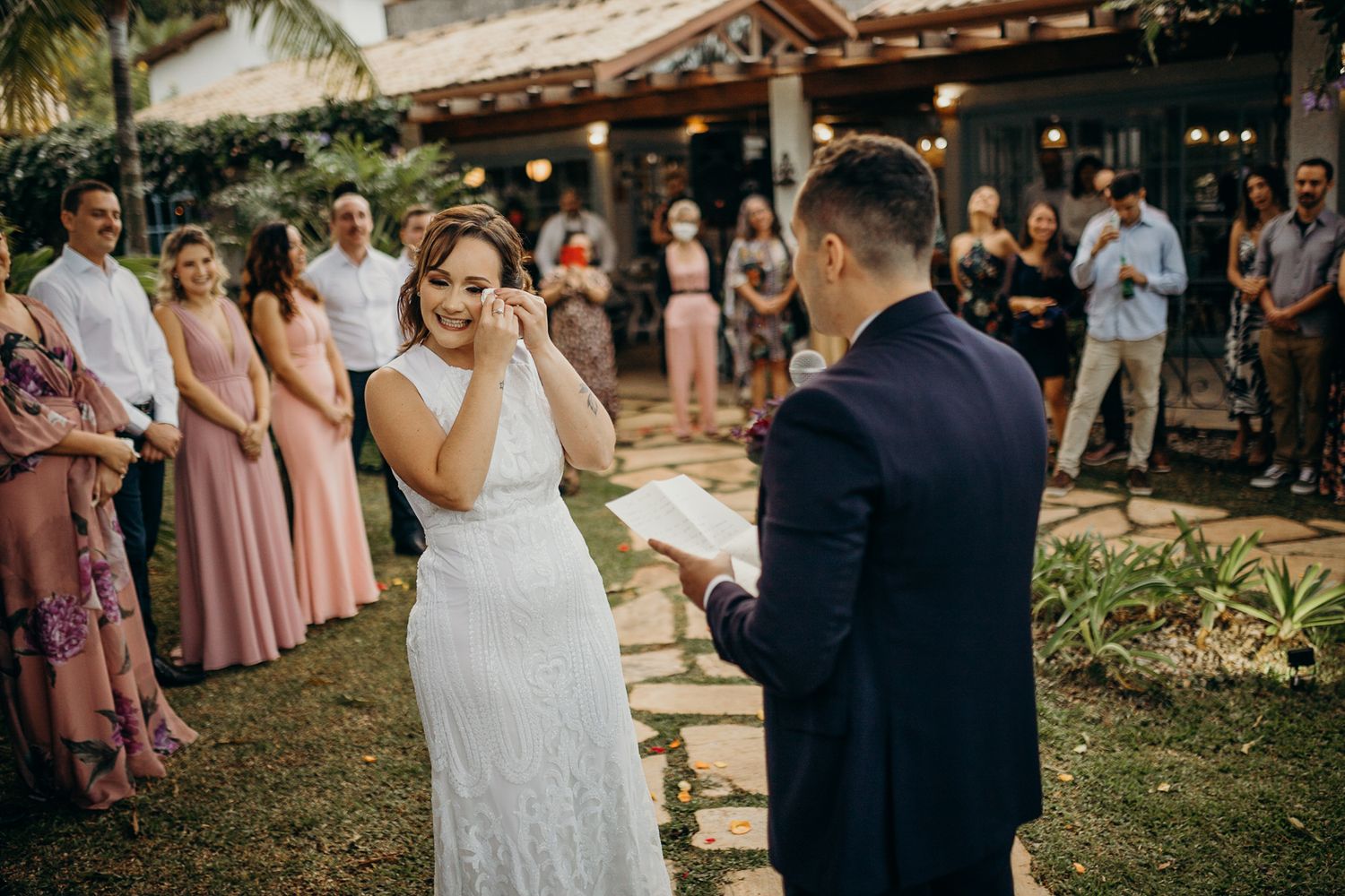 An outdoor wedding ceremony unfolds on a stone pathway with guests watching from a landscaped garden.
