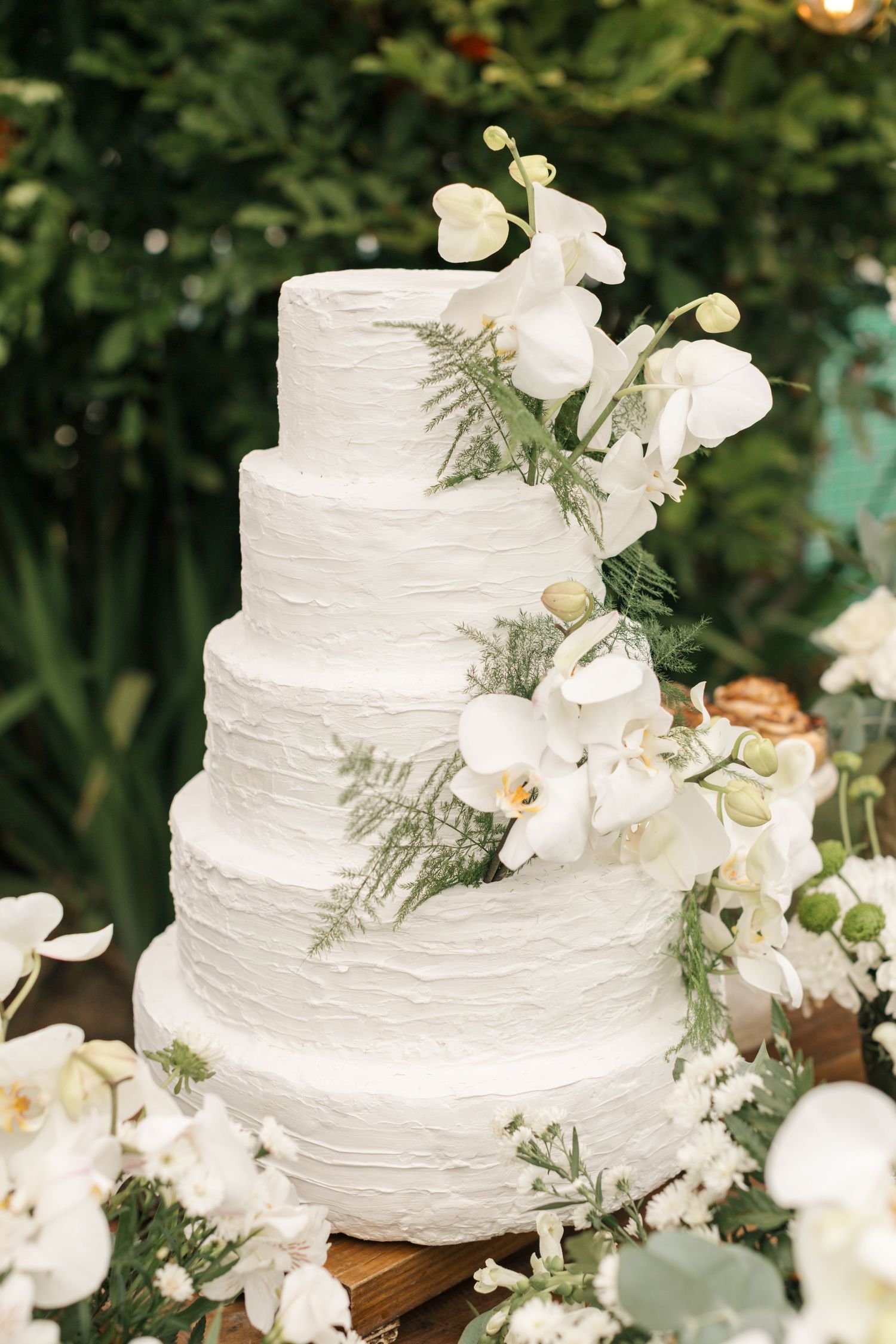 Multi-tiered white wedding cake decorated with fresh orchids and greenery against a garden backdrop.