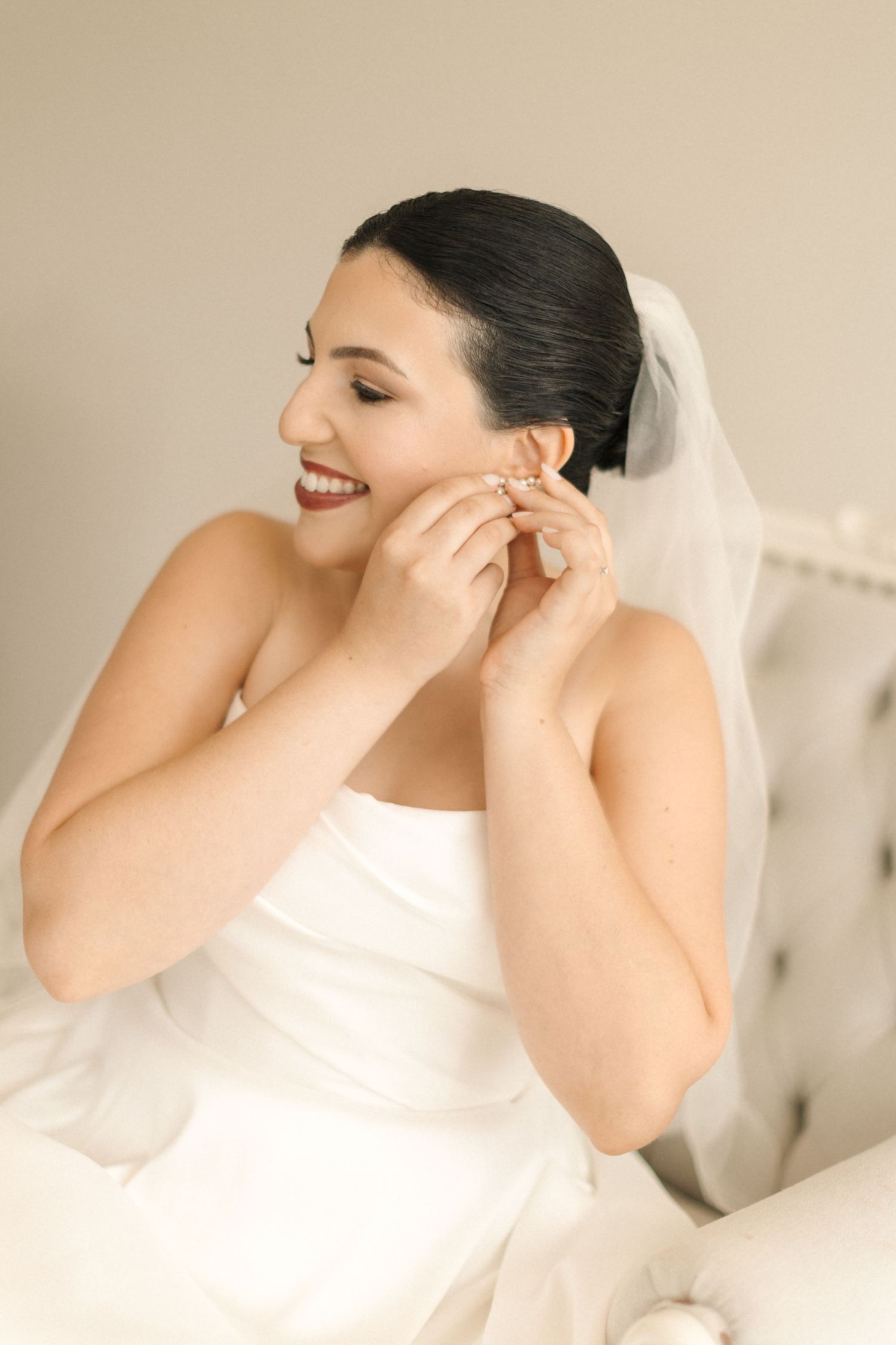 A bride wearing a white wedding dress laughs while putting on earrings during wedding preparations.