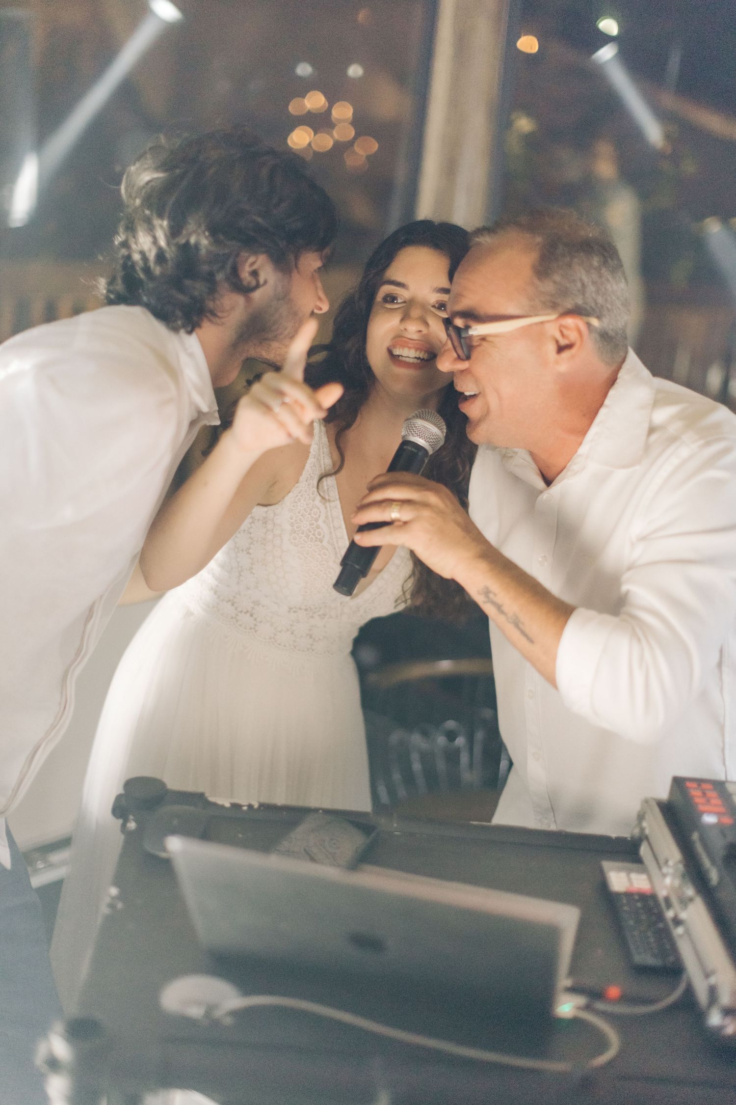 Wedding party members sing karaoke together at a reception with a DJ setup and festive lighting.