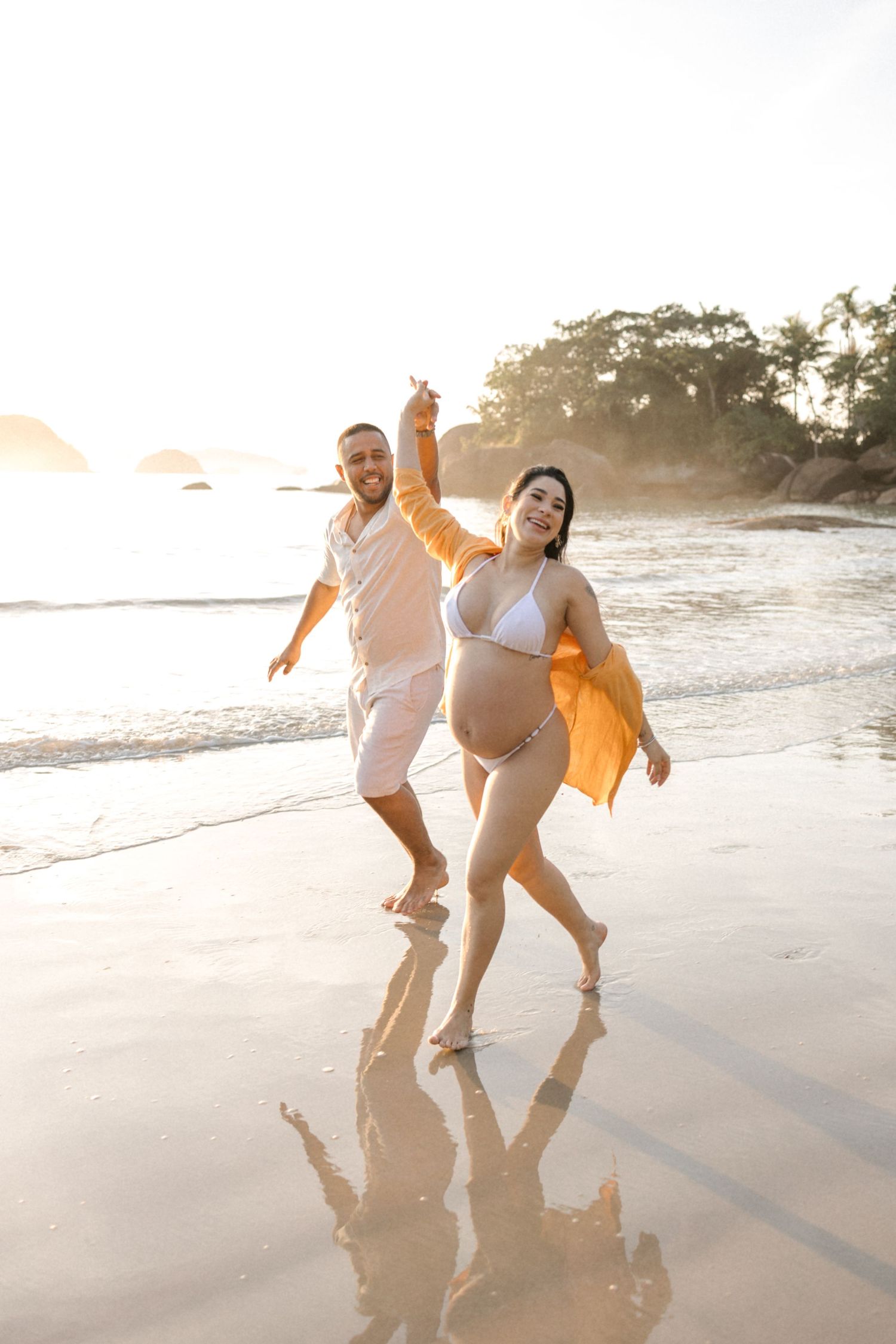 Couple in white clothing dances joyfully on wet sand during golden hour at the beach.