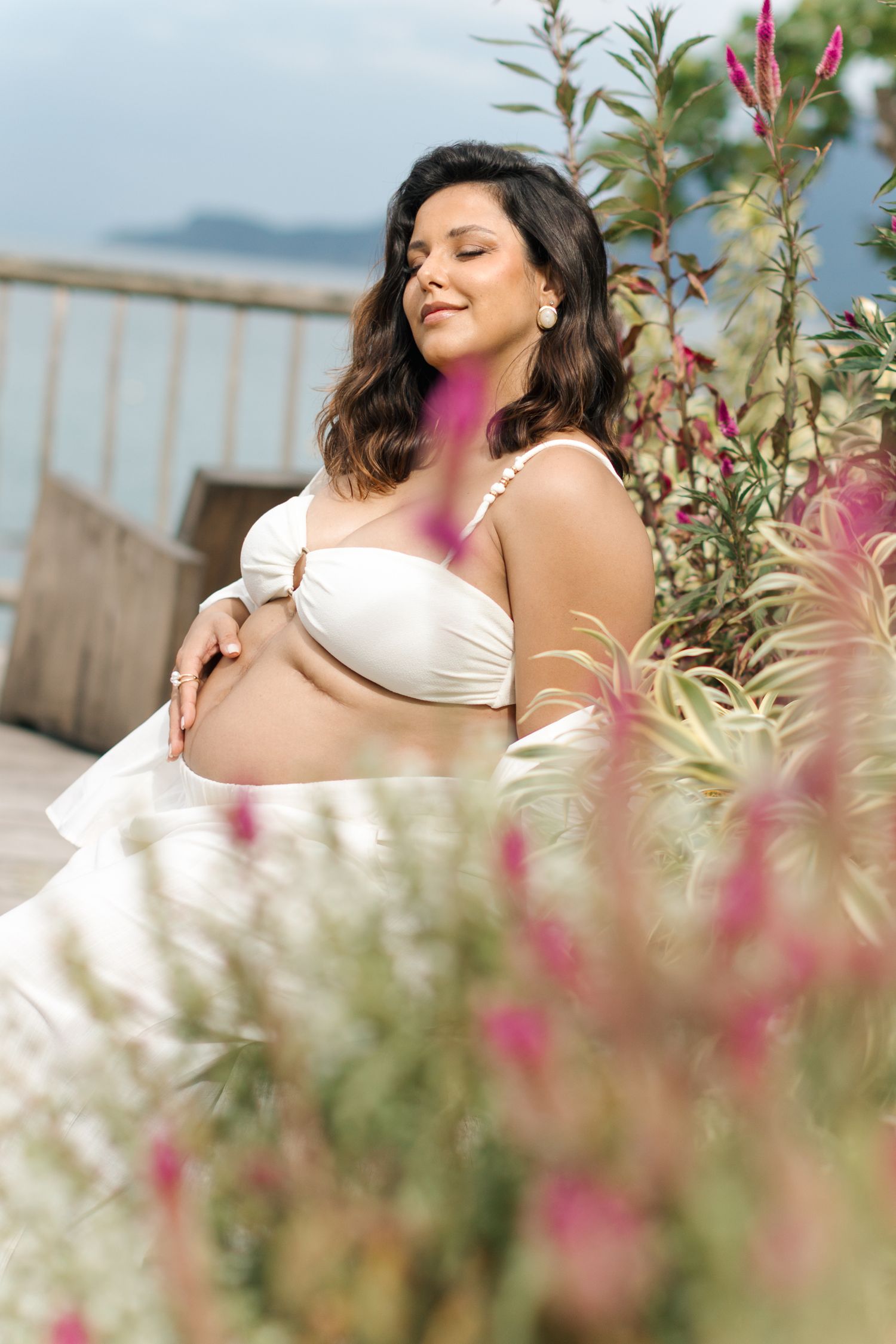 A woman in a white dress poses outdoors among flowering plants and greenery with a scenic background.