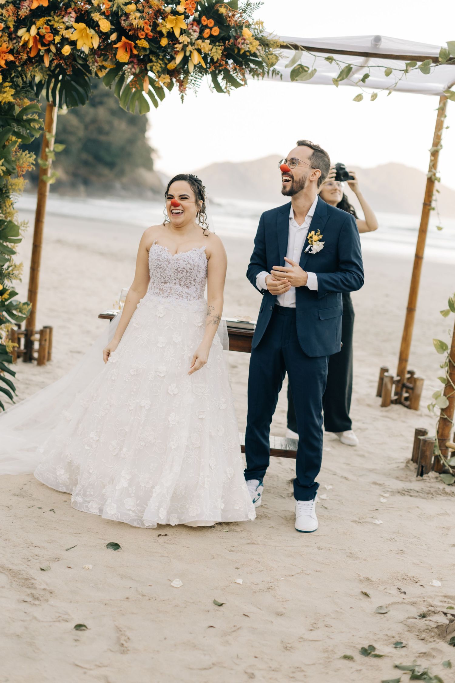 A couple celebrates on a sandy beach with laughter beneath sunlit floral wedding decor.