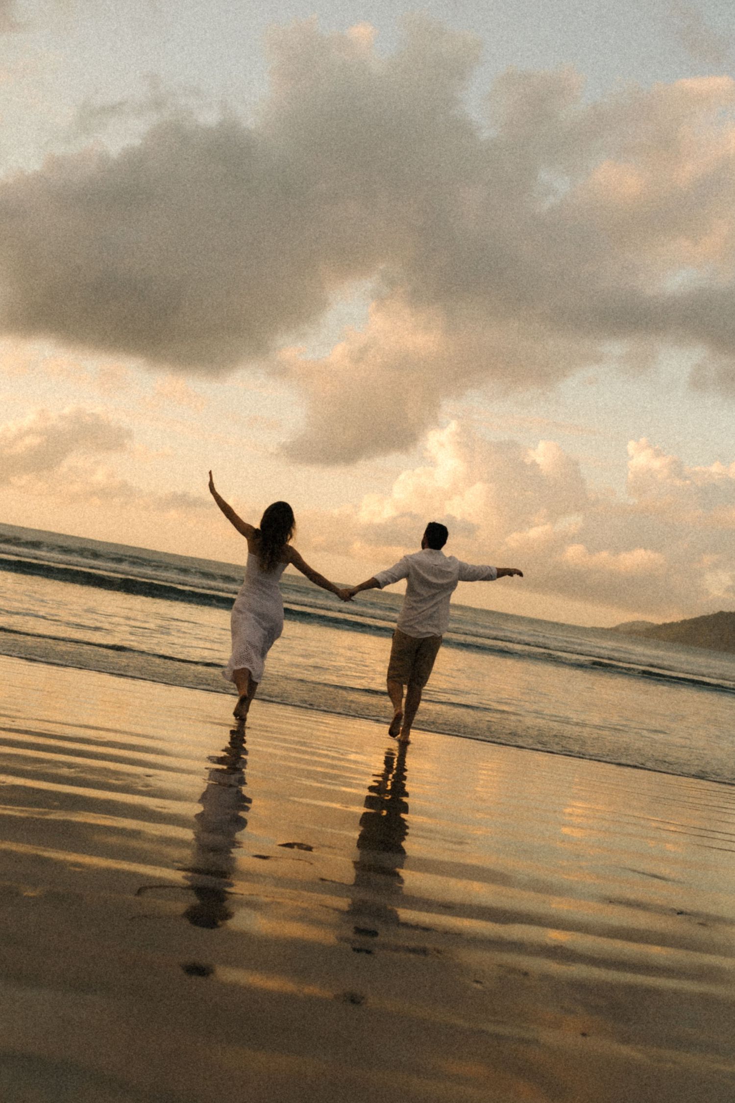 Silhouettes of a couple holding hands and walking on a beach at sunset with golden clouds.