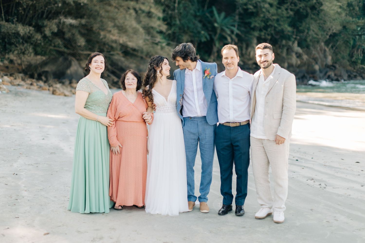 Wedding party and guests in pastel attire pose together on a scenic beach.