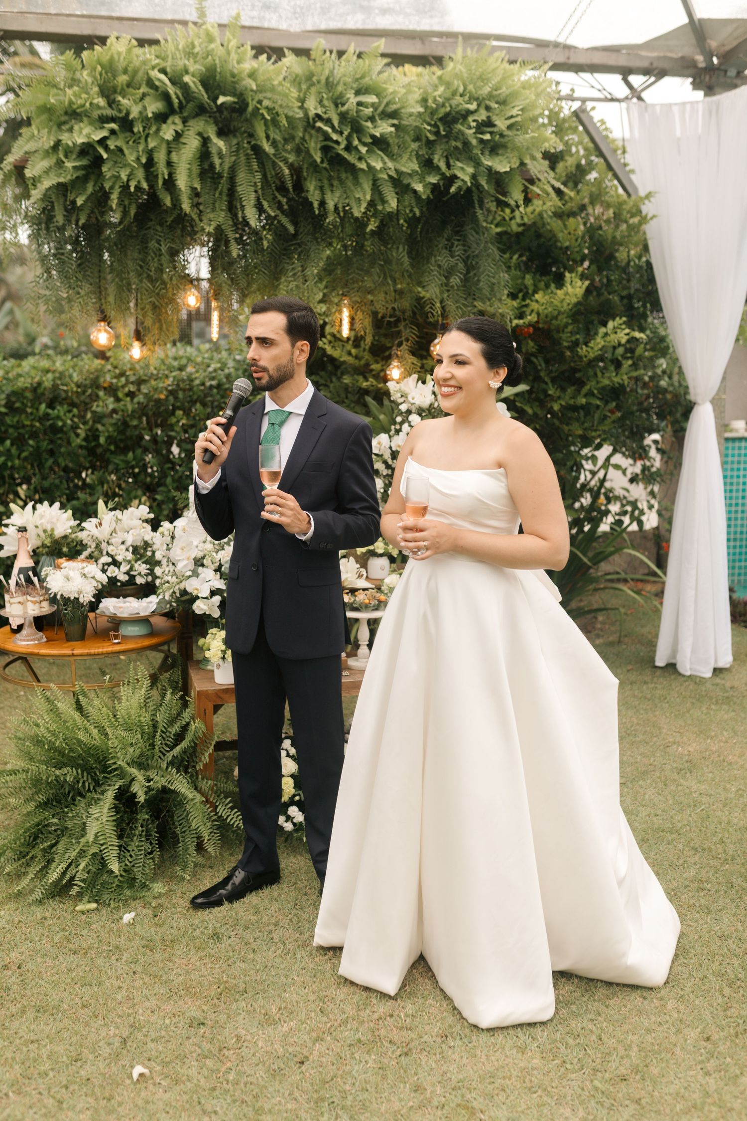 Wedding couple making a speech under hanging greenery and string lights.